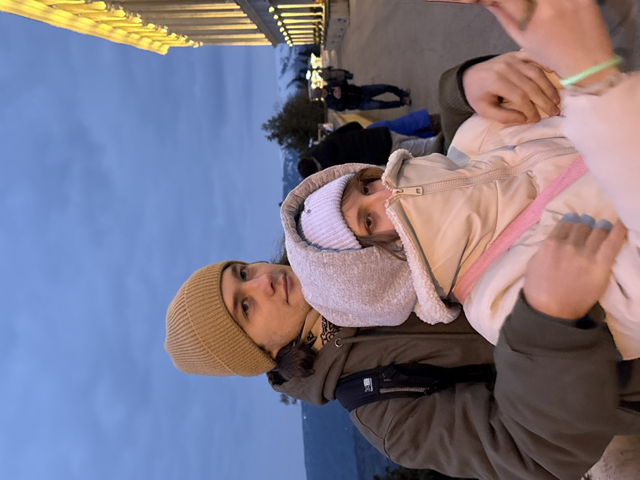 An evening scene in Tbilisi, Georgia, captures two individuals in the foreground on a paved walkway near the Parliament of Georgia building. The sky is dark blue with visible clouds, indicating dusk or early night.
In the foreground, a young adult male stands behind a young adult female. The male wears a light brown ribbed beanie, a dark olive-green hooded jacket, and a patterned scarf visible around his neck. His dark hair extends past his ears. His right arm is wrapped around the female's shoulder, and his left hand rests on her front, near her left arm, holding her. He looks slightly to his right. A dark backpack with a white logo is visible on his left shoulder strap.
The female wears a light-colored, possibly off-white, puffy winter coat with a high zippered collar that obscures her chin and mouth. She has a light purple ribbed beanie underneath a light grey, sherpa-lined hood that frames her face. Her light brown hair is visible around her face. Her eyes are visible, looking downwards and to her right. A pink strap, possibly from a bag or phone sling, crosses her chest. Her left hand, with light blue painted fingernails and a bright green wristband, holds a white smartphone on the right side of the frame, partially obscured.
In the mid-ground, several other indistinct figures are visible on the walkway. One man in a dark jacket and blue pants walks towards the right. Further back, a child in blue is partially visible.
The environment includes a wide, paved terrace or walkway. To the right, a large building with classical-style columns is brightly illuminated with yellow light, consistent with the facade of the Parliament of Georgia building on Rustaveli Avenue. In the distance, under the dark sky, faint outlines of hills or mountains with patches of snow are visible.