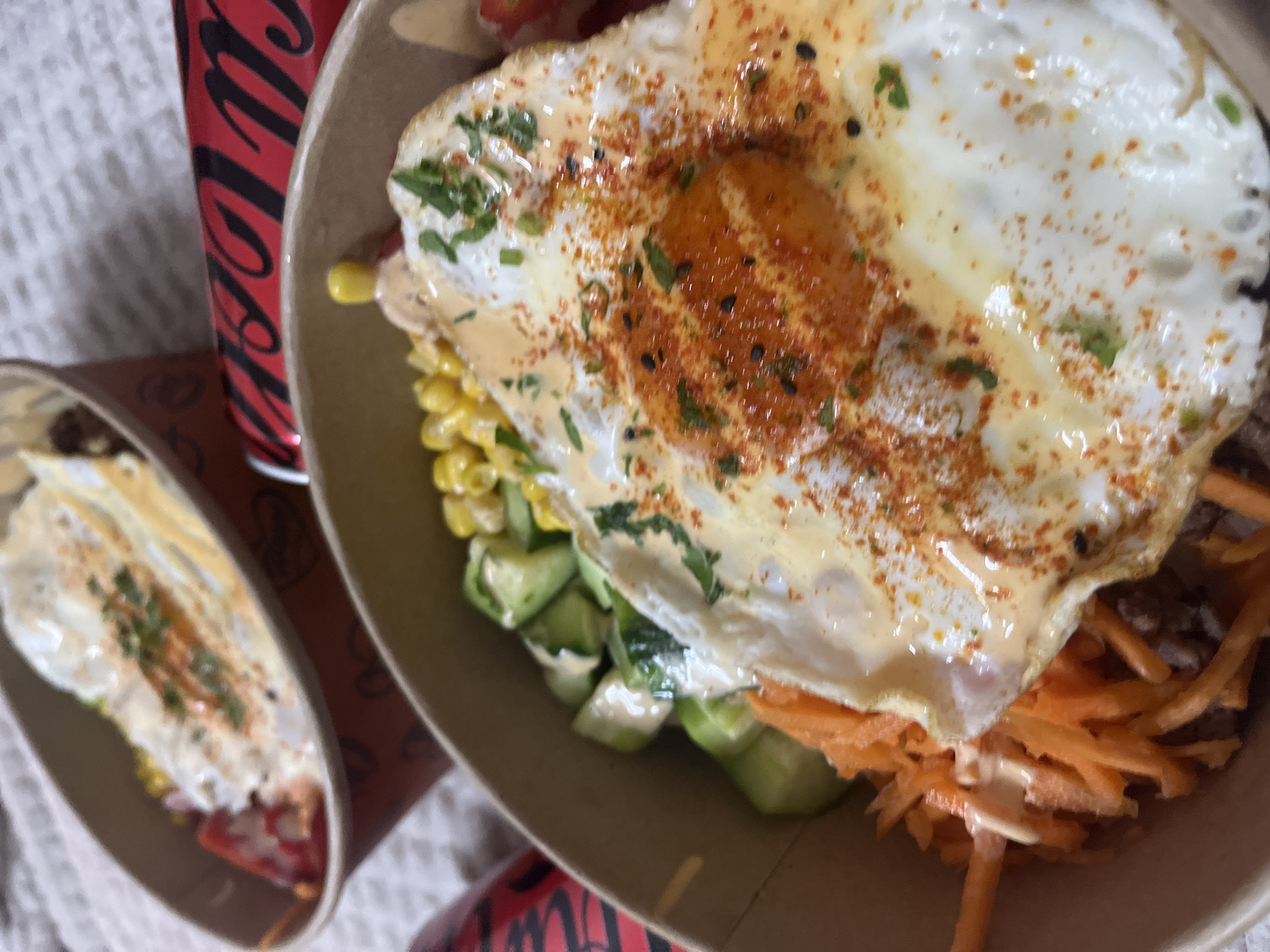 The image presents a close-up view of a vibrant and appetizing meal served in a brown, seemingly disposable, bowl. The central feature of the dish is a perfectly cooked fried egg, likely sunny-side-up, with a slightly runny yolk, generously sprinkled with red spices (possibly paprika or chili powder), black sesame seeds, and chopped green herbs. Beneath the egg, a colorful array of ingredients is visible, including bright yellow corn kernels, diced green vegetables (appearing to be cucumber), and shredded orange carrots. A creamy, light-colored sauce is drizzled over the entire meal.
To the left, a smaller, similar meal in another out-of-focus bowl is partially visible. A red Coca-Cola can, featuring its distinct black script logo, stands partially in the background on the left side, indicating a beverage accompanying the meal. The setting appears casual, with a textured light-colored surface (perhaps a tablecloth or blanket) and a darker, patterned surface underneath the bowls. The photograph captures the meal ready for consumption, suggesting a dining moment. No people are visible. The visible text is the "Coca-Cola" logo on the can.