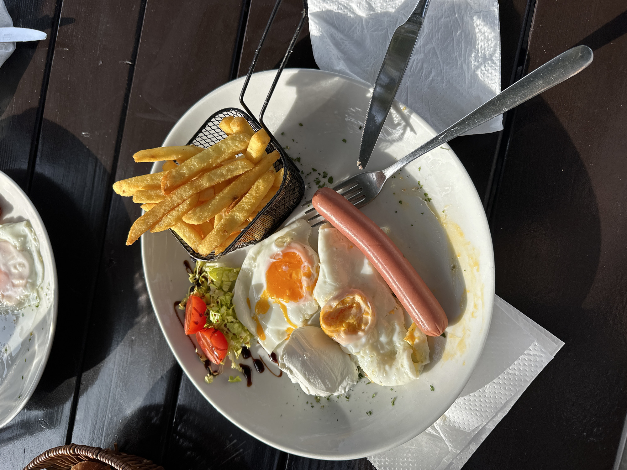 The image shows a white plate on a dark brown wooden table. The plate contains a meal consisting of French fries in a small black wire basket, two fried eggs, a sausage, a small salad of lettuce and tomatoes with balsamic glaze, and a piece of white cheese. A fork and knife lie on the plate, resting on a white napkin. Another plate, partially visible, with a similar meal, is in the lower left corner. A wicker basket peeks into the bottom left corner of the image. The scene suggests an outdoor dining setting, likely in Bar, Montenegro, judging by the context. The lighting indicates it is daytime. There is no evidence of people in the frame.