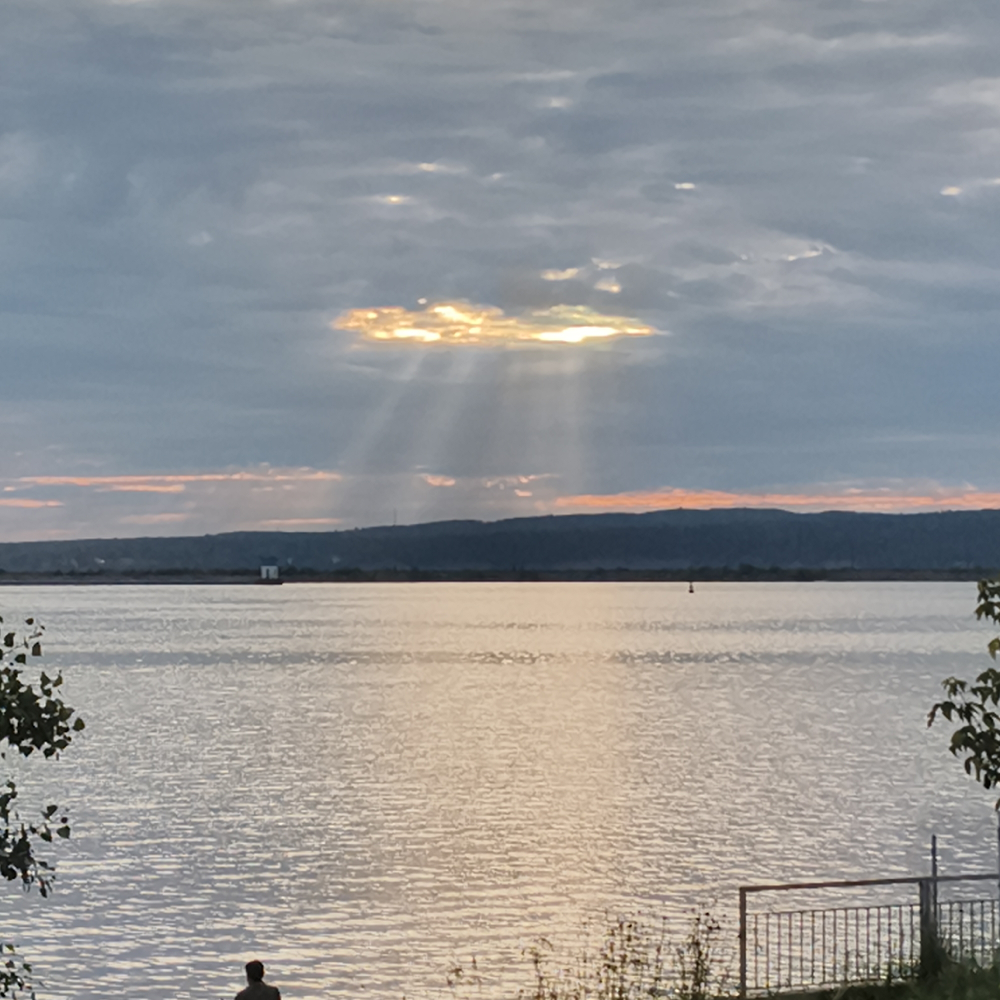 A wide shot captures a serene lake scene in Chaikovsky, Russia, at sunset. The sky is a canvas of dramatic clouds, with a prominent break allowing golden sunbeams to pierce through, illuminating the water below. The sun's rays fan out, creating a ethereal glow. Hues of orange and pink streak the horizon, suggesting the fading light of day.
The lake itself is vast and calm, reflecting the soft light and the colors of the sky. In the distance, a dark, silhouetted treeline forms the far bank of the lake, with a small, box-like structure visible on the shore. Further out in the water, a single buoy marks the expanse.
In the foreground, a person sits facing the water, their back to the camera, appearing contemplative or at peace. To the right, a simple metal fence runs along the bank, with some greenery and foliage in the lower corners of the frame. The overall mood is tranquil and reflective, emphasizing the beauty of nature at dusk.