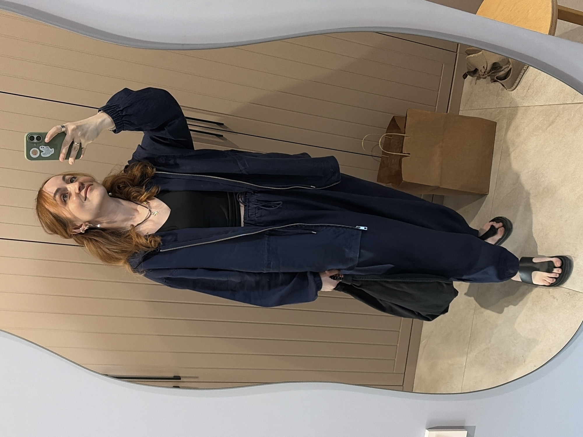 A woman is taking a selfie in a mirror. She is wearing a black top, a navy blue jacket, and navy blue pants. She is also wearing black sandals. Her hair is reddish-brown and she has a silver necklace and earrings. She is holding a green phone in her left hand. There is a brown paper bag on the floor next to her. In the background, there is a beige wall and a wooden chair. The location is inside a store in the city of Chaikovsky, Russia.
