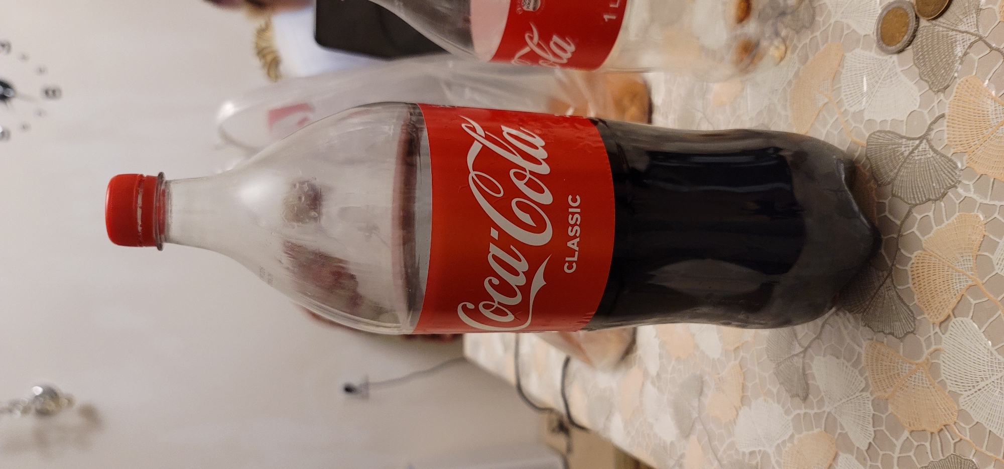 The image shows a Coca-Cola bottle with a red cap, filled with dark liquid. The bottle has a label that says "Coca-Cola CLASSIC" in white lettering on a red background. The bottle is standing on a patterned tablecloth. In the background, another partially visible bottle and some blurry objects can be seen. The scene appears to be indoors, possibly in a domestic setting. There is no indication of the location being in Ganja City, Azerbaijan.