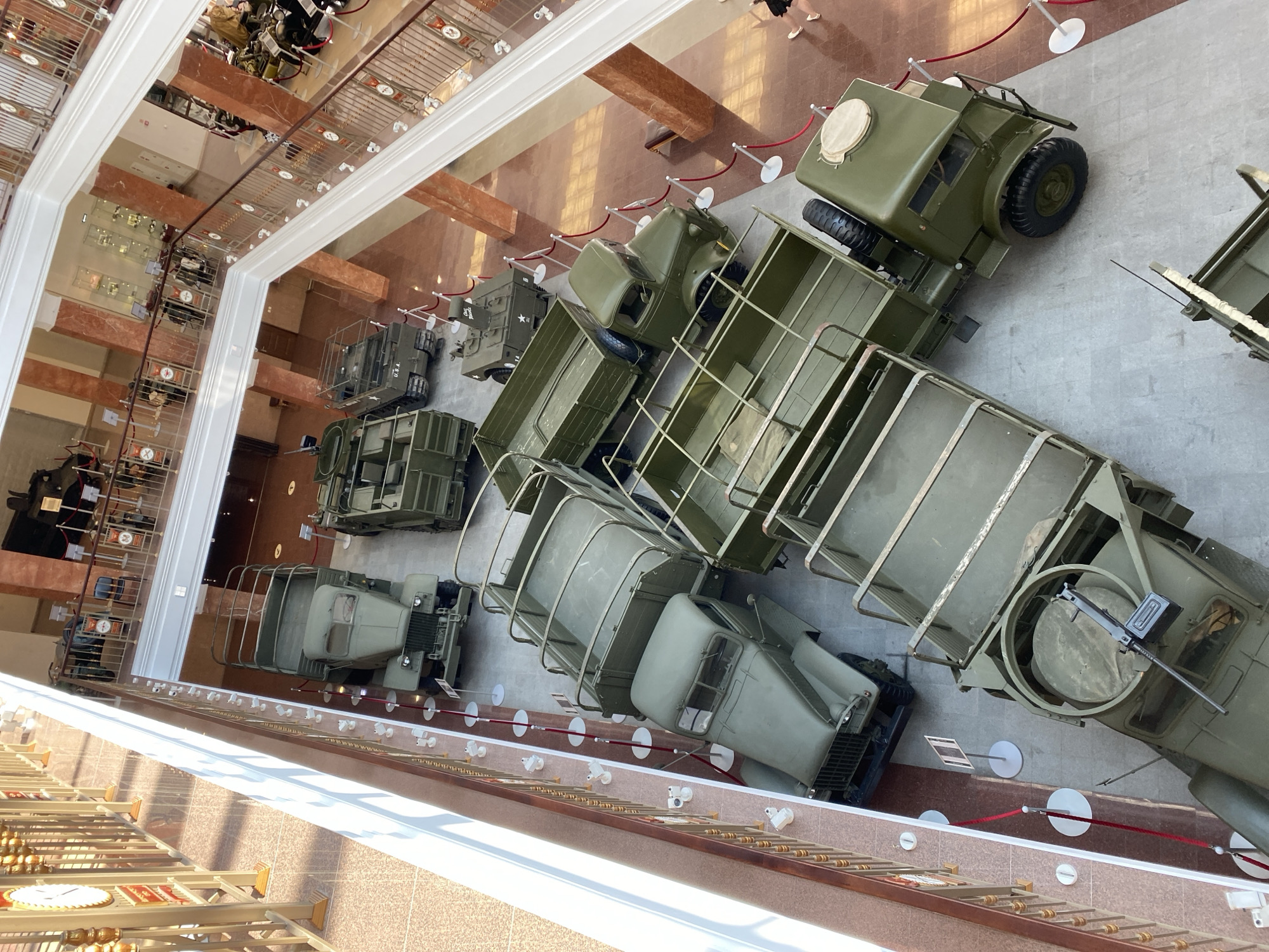 An overhead, high-angle shot showcases the interior of a museum, likely the Military Technology Museum in Verkhnyaya Pyshma, Russia, as evidenced by the collection of military vehicles. The museum floor is a light grey, tiled surface. Dominating the scene are several olive-drab military trucks, arranged in rows and on multiple levels of the building. The trucks appear to be from various eras, some with open cargo beds and others enclosed cabins. Some trucks are equipped with what appear to be machine guns mounted on their turrets.
On the upper levels of the museum, visible through open walkways and railings, are more exhibits, including what seem to be other vehicles and display cases. The architecture features polished red marble columns and ornate golden railings. Natural light streams in from the left, casting long shadows across the floor and the vehicles. Red velvet ropes on white stanchions cordon off the exhibits. Two individuals are visible on the far right, one wearing dark clothing and the other in white shorts and a black top, walking on a lower level of the museum. A large clock is partially visible on the far left of the upper level.