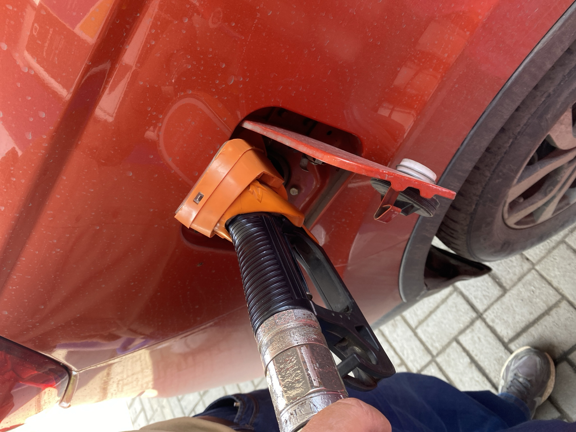 A person is at a gas station in Novokuznetsk, Russia, filling their orange car with fuel. The nozzle of the gas pump is inserted into the car's fuel tank. The car is dirty with water droplets on its surface. The ground is paved with light-colored bricks. The person is wearing blue jeans and gray sneakers. The scene is captured in a close-up, slightly angled shot from the right side of the car.