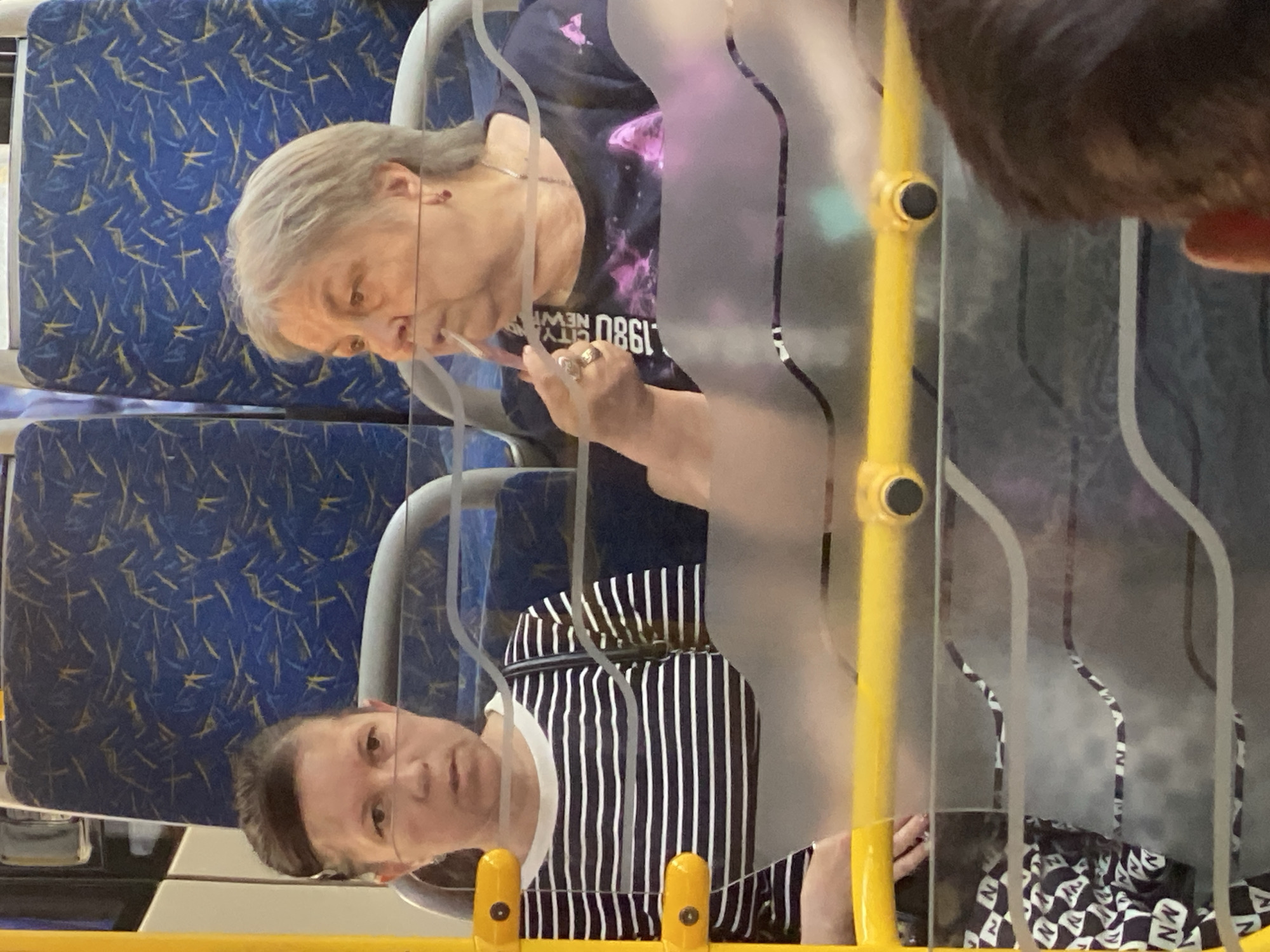 The image shows two women seated on a bus with blue and yellow patterned seats. A clear plastic partition separates them from the foreground. The woman on the left is wearing a black and white striped shirt and has her hair pulled back. The woman on the right has gray hair and is wearing a dark t-shirt with a pink graphic and the text "1980 CITY NEW Y..." visible. She is holding a pink object to her mouth. The bus appears to be in motion, as evidenced by the blurred reflections on the partition and the slight motion blur in the background. The location is the city of Novokuznetsk, Russia.