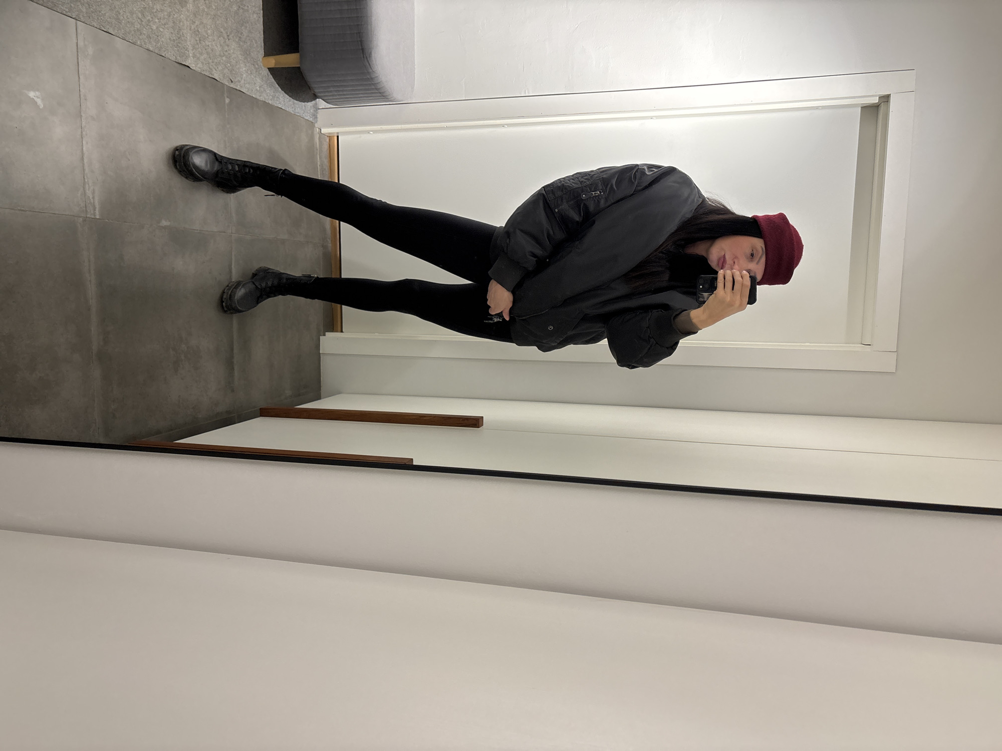 A person in a mirror selfie is positioned in front of a white wall. The person has dark hair, is wearing a dark bomber jacket, black pants, black boots and a burgundy beanie. The floor is grey tile. A grey piece of furniture is visible. The person is holding a black phone. The location cannot be identified.