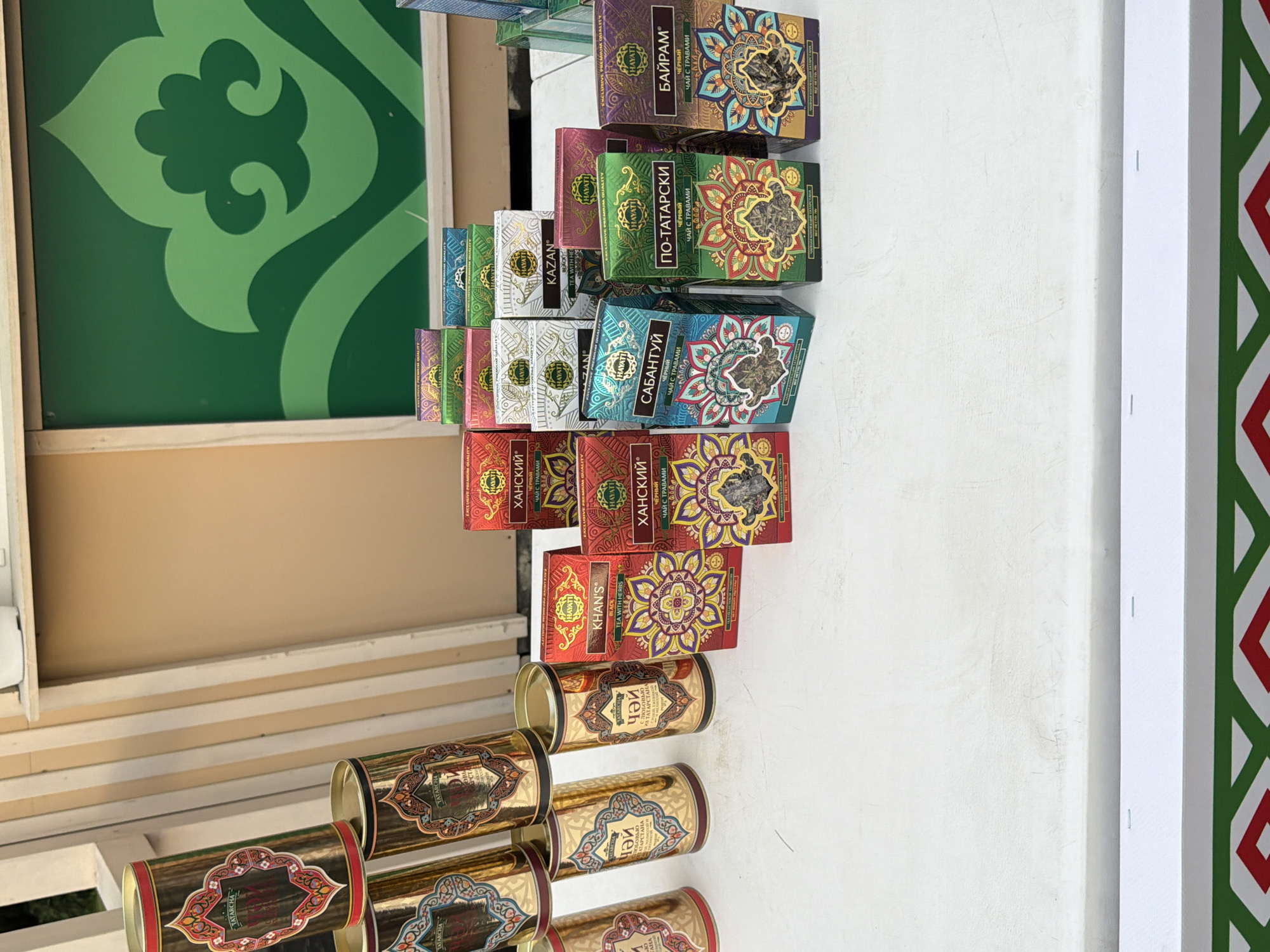 A close-up, eye-level shot shows a collection of cylindrical tea tins and rectangular tea boxes arranged on a white surface, likely a table. The tea containers are predominantly gold with ornate, colorful patterns in red, blue, green, and white, featuring intricate floral and geometric designs. Some tins have visible labels with Cyrillic text and the brand name "TATARCHA". The tea boxes are stacked in rows and are also brightly colored with similar decorative motifs. Text on the boxes includes brand names like "KHAN'S", "HAYATI", and "KAZAN", along with descriptions in Cyrillic. In the background, a portion of a beige wall and a vertical structure composed of light-colored planks are visible. To the right of the wall, a large green graphic with a swirling design, reminiscent of traditional patterns, is displayed on a panel. The lighting suggests an outdoor or well-lit indoor setting.