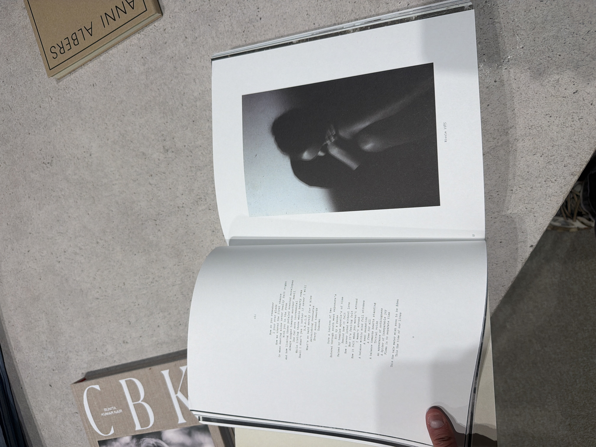 The image shows an open book lying on a light grey surface. The book contains a black and white photograph on the top page, labeled "Nicole 1985." The bottom page contains poetry. Two other books are partially visible near the primary book: one labeled "ANNI ALBERS" and another with "C-BK SUNITA KUMAR NAIR" visible on its cover. A finger is visible in the lower right corner of the frame. The location is not specified in the image.