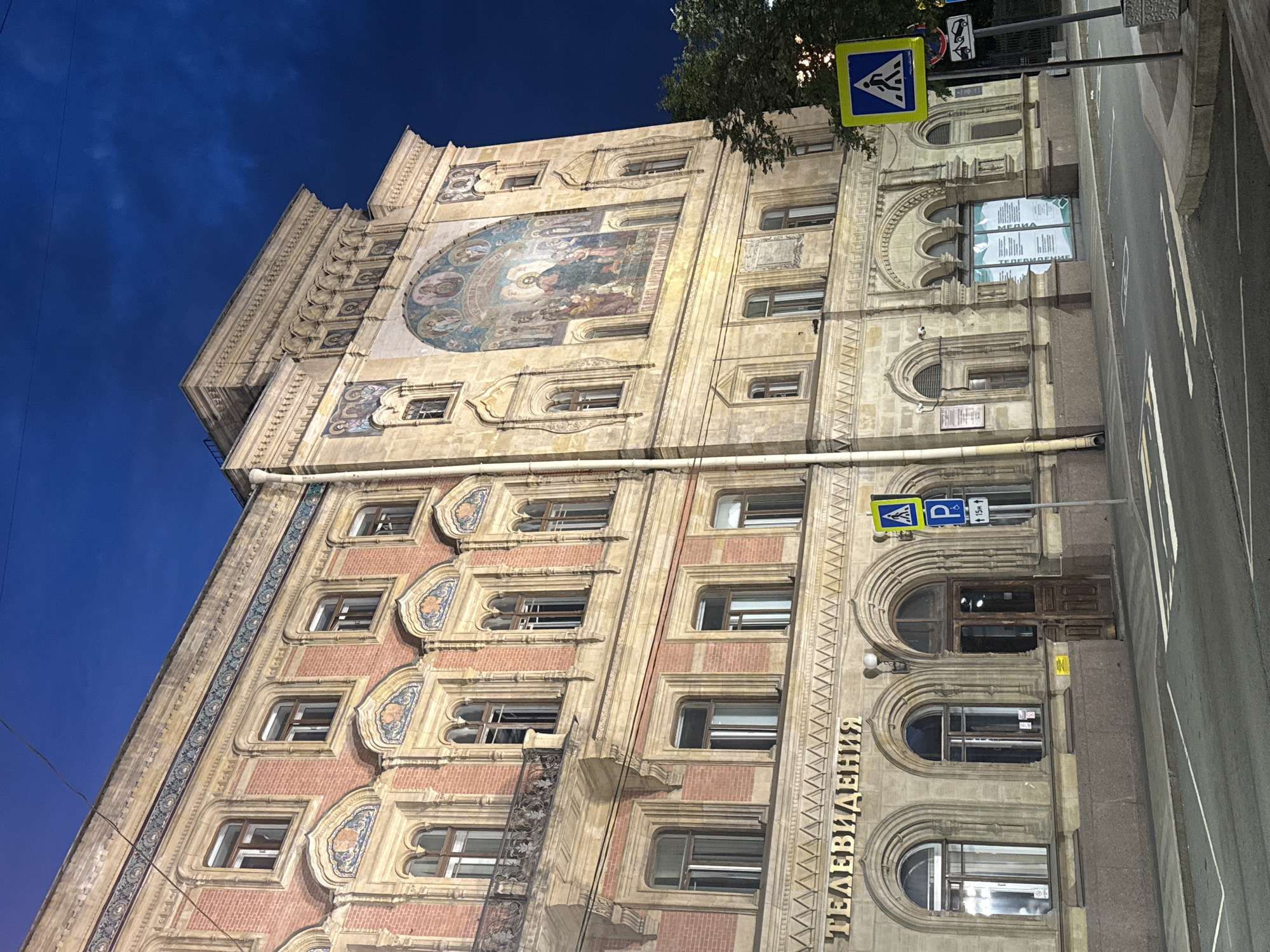The image captures the facade of a grand, historic building in Saint Petersburg, Russia, under a deep blue twilight sky. The building exhibits intricate architectural details, including brickwork, ornate window frames with arched tops, and decorative friezes. A prominent fresco, depicting religious figures, adorns the upper portion of the building's facade. The ground floor features several arched doorways and windows. A sign with Cyrillic lettering, reading "ТЕЛЕВИДЕНИЯ" (Television), is visible above a set of arched entrances. Across the street, a pedestrian crossing is marked, and a pedestrian crossing sign is posted. The street is paved, and there are visible lane markings and parking spaces. The overall scene suggests a quiet urban evening in a historically rich area of the city.