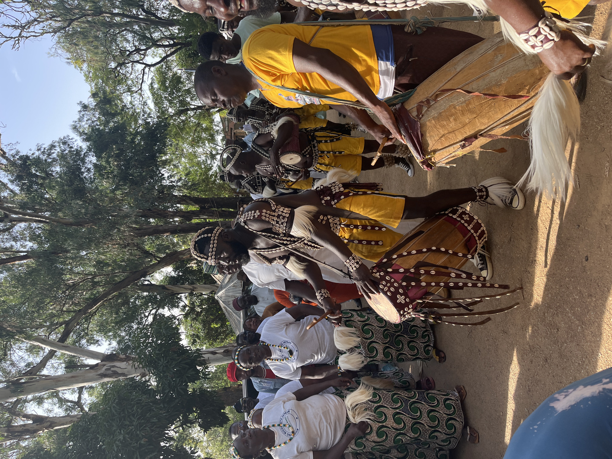 In the city of Jos, Nigeria, a group of people are gathered outdoors, with many individuals dressed in traditional attire. The foreground is dominated by a man in the center, wearing a yellow tunic and brown shorts, actively playing a large drum with drumsticks. To his left, another man in a similar yellow outfit also plays a drum. Several women are visible to the left, wearing white t-shirts and patterned skirts, some holding smaller drums or decorative items. The attire of the performers includes intricate beadwork, shells, and white feathers. The environment is characterized by dirt ground and a backdrop of tall trees with lush green foliage, under a bright, clear sky. A tent or canopy is partially visible in the background, suggesting a gathering or event. The overall scene depicts a cultural performance or celebration.