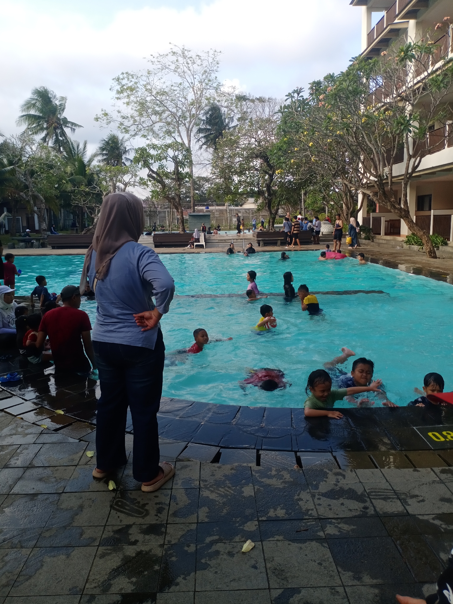 This is an outdoor daytime, eye-level, vertical shot of a swimming pool area in Desa Pagandaran, Indonesia. A woman wearing a light blue shirt and dark pants, with a brown headscarf, stands facing away from the camera on the tiled pool deck, her right hand on her hip. She appears to be supervising.
The pool is filled with clear turquoise water and populated by numerous children of various ages, some playing, some swimming. A few adults are visible in and around the pool. In the background, there is a multi-story building with balconies and an outdoor area with trees and palm trees. The sky is partially cloudy. The pool deck is wet and has some fallen leaves. One child in the foreground is wearing a green shirt and is reaching out towards the camera. Another child, wearing a blue and black patterned shirt, is also in the water near the front. A red object, possibly a pool toy, is visible in the lower right corner. The overall scene depicts a recreational setting, likely a hotel or resort, with people enjoying a day at the pool.