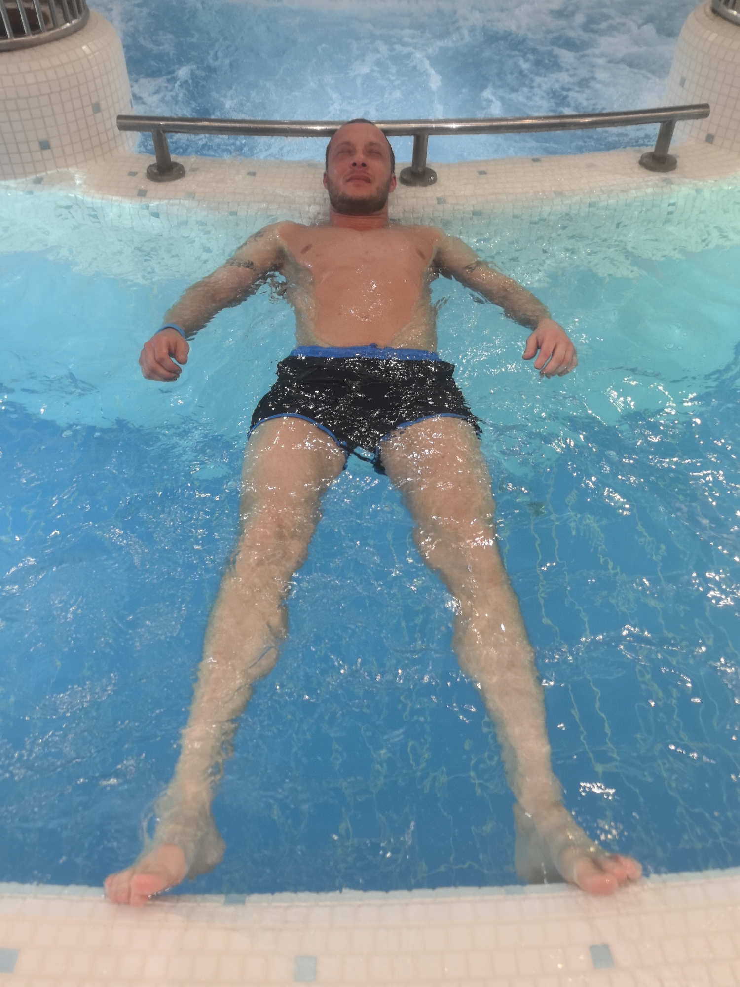 A man is floating in a pool or hot tub, lying on his back with his arms and legs extended. He is wearing black swim trunks with a blue waistband. He has short hair and a light beard. The water is a bright blue color, and the pool is surrounded by white and light blue tile. A metal railing runs along the edge of the pool. The location is implied to be in the city of Nizhny Novgorod, Russia.