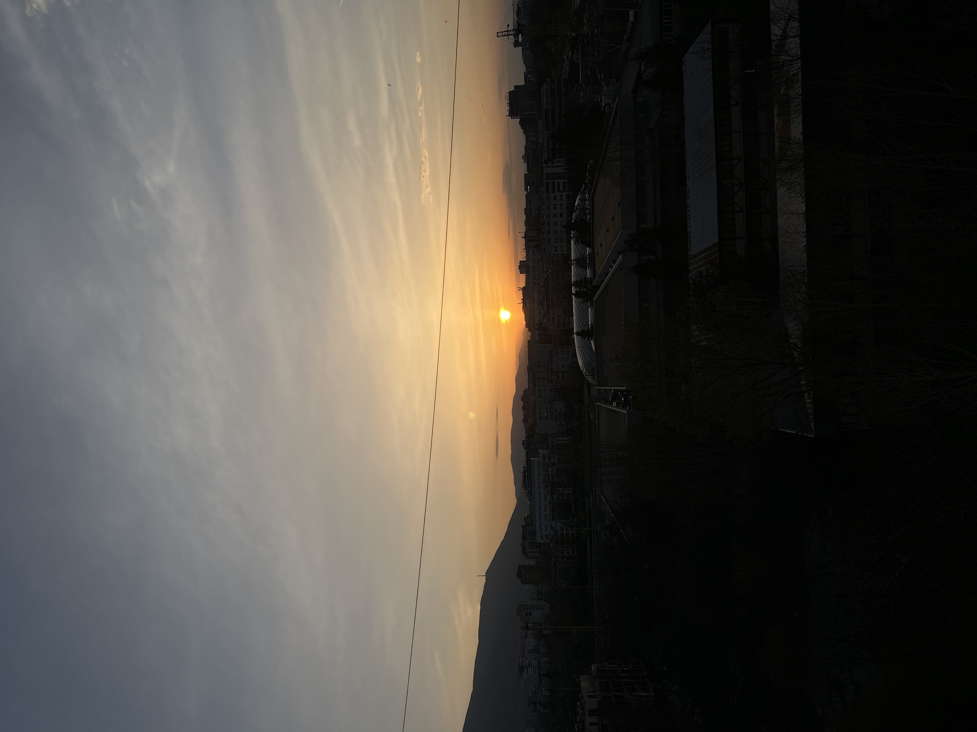 The image shows a sunset over Sofia, Bulgaria. The sun is setting behind the Vitosha mountain, casting an orange glow on the sky. A city skyline is visible, with buildings in silhouette. There are power lines in the left side of the image. The sky is a mix of gray and orange. Part of the image is dark because of the direction of sunlight and location.