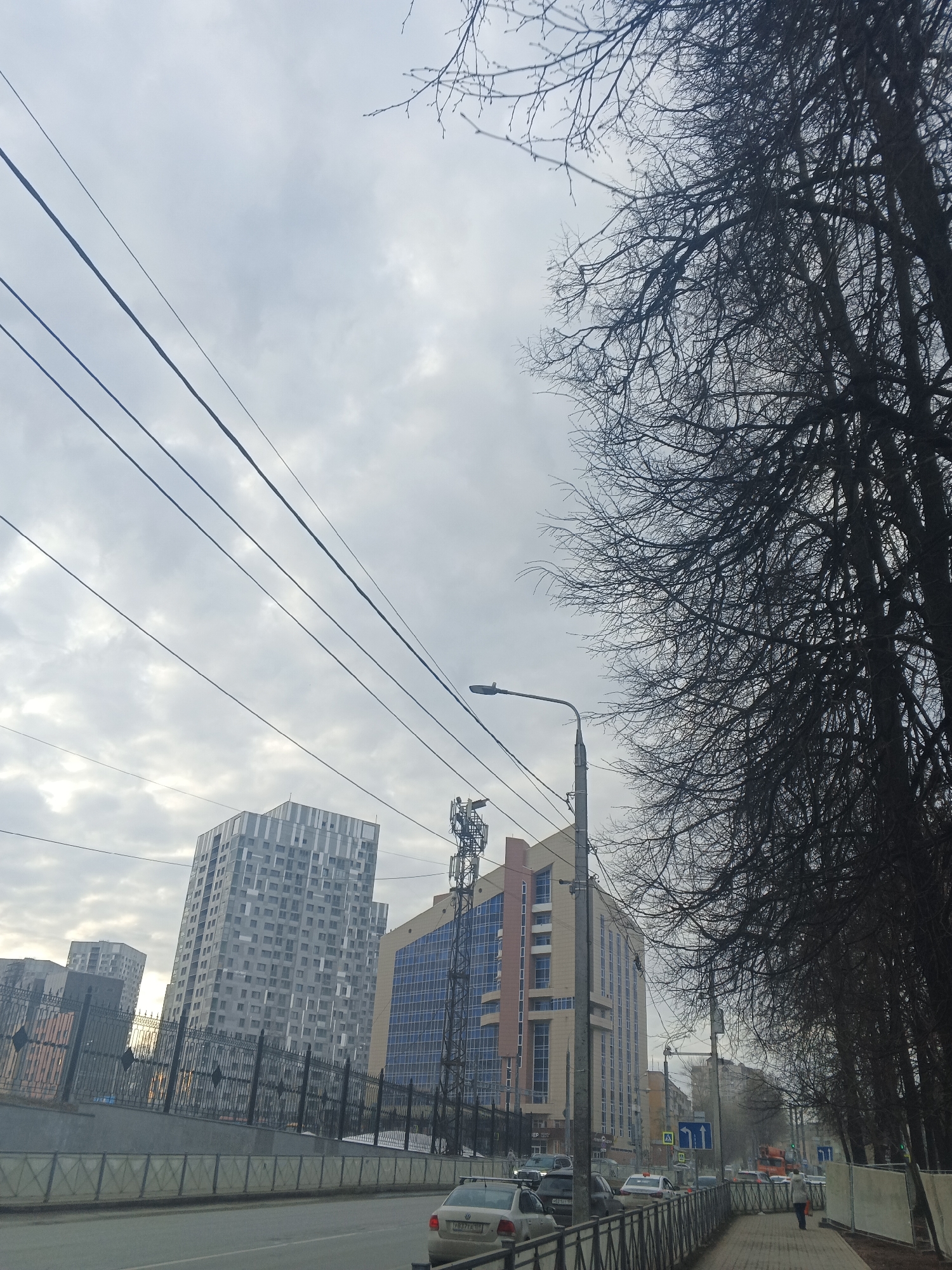 The image depicts a street scene in Perm, Russia on an overcast day. In the foreground, a grey car is visible, with a metal fence separating the road from the sidewalk. A few other cars can be seen further down the road. A pedestrian is walking on the sidewalk. Several buildings are visible in the background, including a tall grey and white building with multiple windows, and a tan and blue building. A utility pole with wires is visible, as well as a street light. Bare trees line the right side of the image, their branches silhouetted against the cloudy sky. Traffic signs are present on the side of the road.