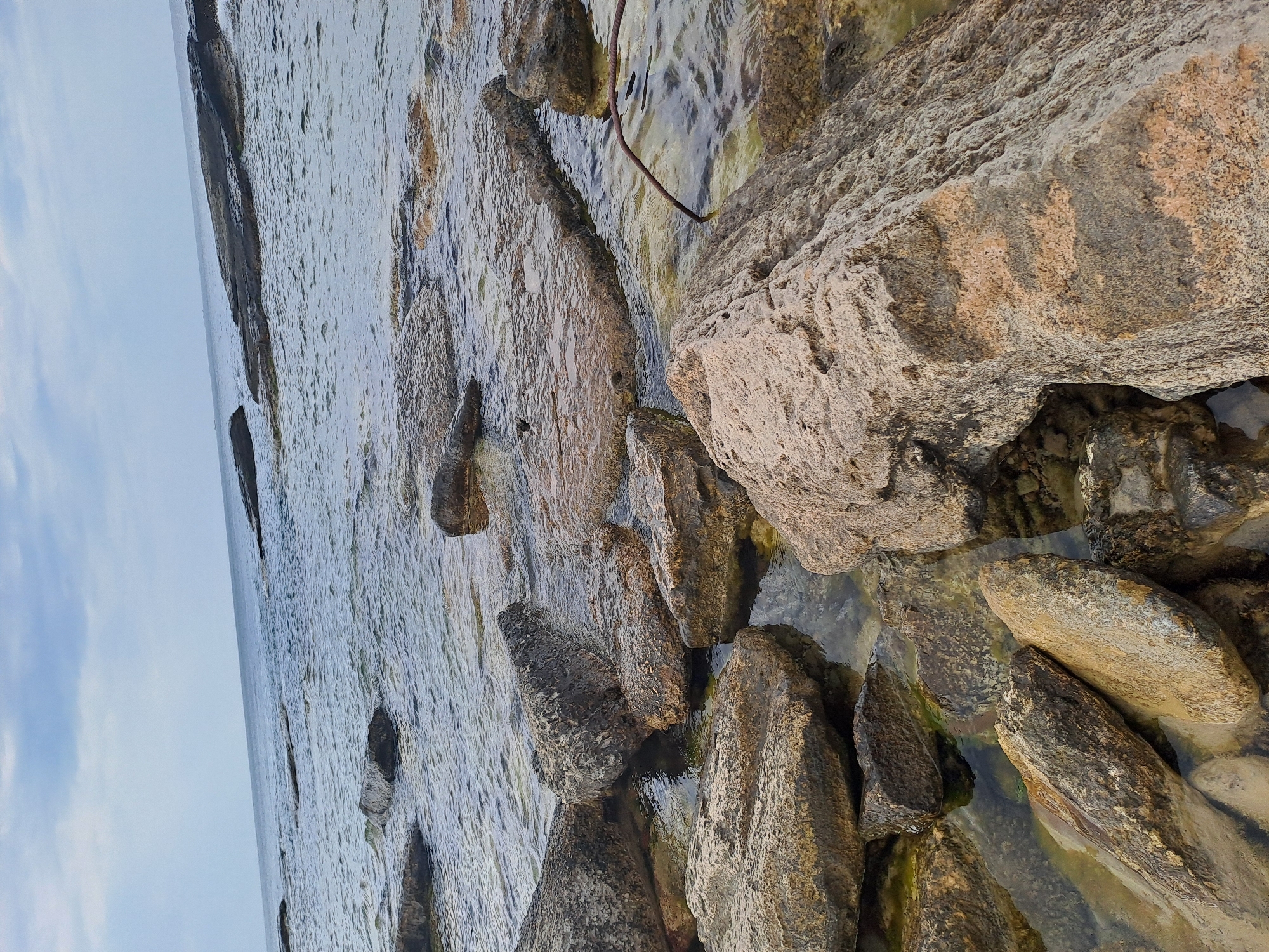 The image depicts a rocky shoreline with the Caspian Sea in the background. The water is clear, allowing a view of the submerged rocks and sand. Large, weathered stones of various shapes and sizes are scattered across the foreground, some partially submerged in the shallow water. The sky is overcast with light gray clouds. There are no people or animals visible in the image. The scene is a natural coastal environment with a slightly weathered and rugged appearance. The location is the coast of Aktau, Kazakhstan, known for its rocky shores and proximity to the Caspian Sea.