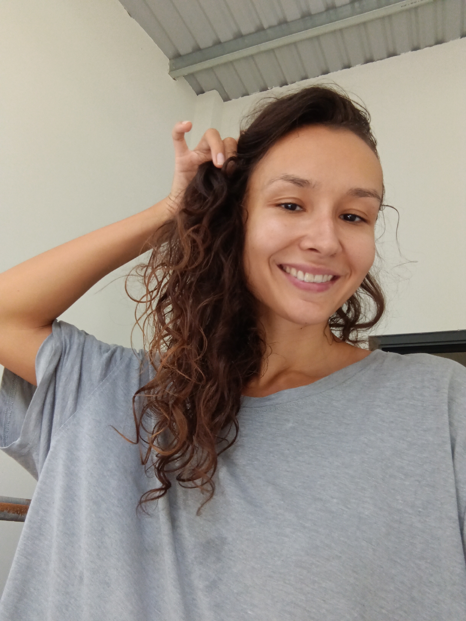 A young woman with long, curly brown hair is shown in a close-up, outdoor shot. She is wearing a grey t-shirt and is looking at the camera with a bright smile. Her right hand is raised to her head, touching her hair. The background is a neutral-colored wall and a corrugated metal roof. The scene appears to be in a casual, possibly residential or commercial area, given the architecture. There are no specific indicators to place the location in Mũi Né, Vietnam.