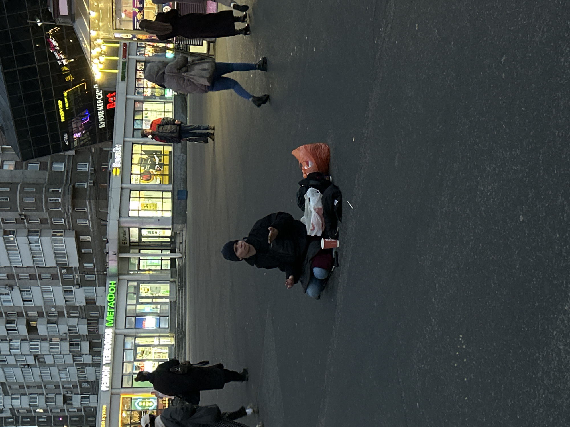 The image captures an urban street scene in Saint Petersburg, Russia, during dusk or early evening. Artificial lights from commercial buildings illuminate the surroundings, suggesting cool weather.
In the midground, a person is kneeling on the dark pavement, dressed in a black jacket, dark pants, and a dark beanie hat. Their hands are slightly open, suggesting they are gesturing or appealing for help. Beside them on the ground are an orange plastic bag, a black backpack, a white plastic bag, and a red and white cup, possibly a coffee cup, with the word "DELICIOUS" partially visible.
Several pedestrians are seen walking in the background. One person wears a light-colored jacket and blue jeans, another a dark jacket with a red backpack, and others are dressed in various dark coats, walking past the kneeling figure.
The background consists of typical city architecture. To the left is a tall residential or mixed-use building with multiple windows and balconies. To the right, commercial buildings line the street, featuring brightly lit storefronts. Visible signs include a red sign with "БУКМЕКЕРСКАЯ" (Bookmaker's), the "Билайн" (Beeline) logo, and a prominent green sign displaying "РЕМОНТ ТЕЛЕФОНОВ МЕГАФОН" (Phone Repair Megafon). A stylized "Burger" sign is also visible. The scene portrays a moment of urban daily life, with the central figure's posture and belongings suggesting an appeal for assistance.