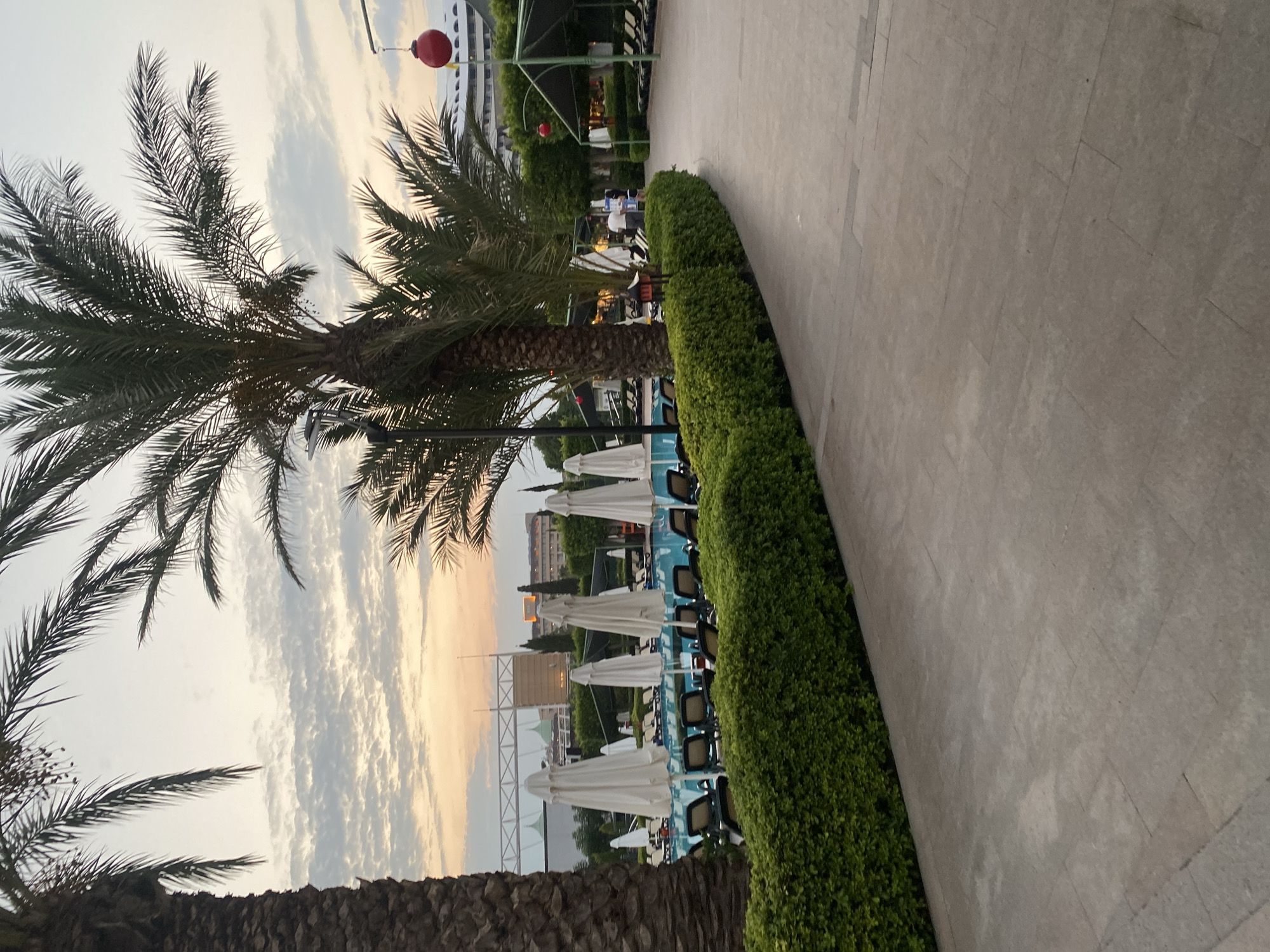 The image depicts a resort area in Aksu, Turkey, near sunset. A paved walkway leads past a neatly trimmed green hedge and a swimming pool area. The pool is lined with lounge chairs and closed white umbrellas, suggesting the end of the day. Several palm trees are visible, framing the scene and casting shadows. In the background, resort buildings and structures, including what appears to be a stage or event area, are partially obscured by trees and the twilight sky. Two red decorative lanterns hang from wires, adding a touch of color to the scene. A few people can be seen in the distance near the pool area, and a street lamp is also present. The sky is filled with soft, layered clouds illuminated by the warm tones of the setting sun.