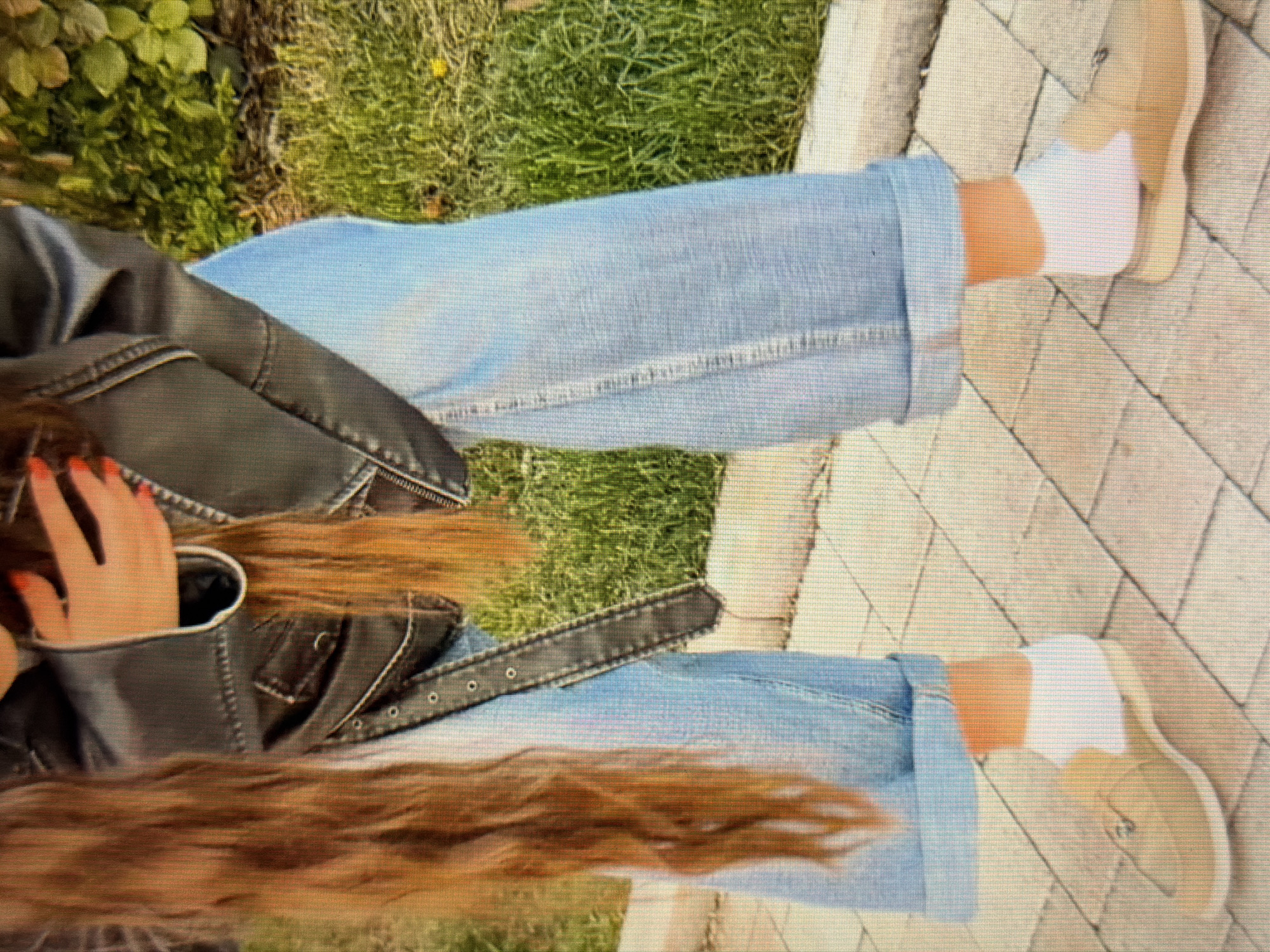 The image shows a close-up view of a person's lower body, from the waist down, standing outdoors on a paved walkway. The person is wearing a black leather jacket, light blue cuffed jeans, white socks, and light brown slip-on shoes. The background consists of green grass and some foliage. The paved walkway is made of rectangular tiles. The image does not contain any information that would allow for the determination of its location in the city of Sirius, Russia.