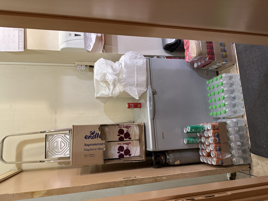 This image captures a storage or backroom area, possibly in a commercial establishment in Nea Erithrea, Greece. The scene is static, showing various supplies neatly organized.
On the left, a silver hand truck or dolly is visible, next to a large cardboard box labeled "endlss Χαρτοπετσέτες Napkins Med." This box is open, revealing two smaller boxes inside, also with the "endless Maga" branding. These boxes are placed on a wooden counter or shelf that spans the foreground of the image.
Towards the right, a silver refrigerator stands against the wall. A crumpled white plastic bag rests on top of it. A red Coca-Cola can is magnetically attached to the wall above the refrigerator. Below the refrigerator, a gray gas cylinder is visible.
To the right of the refrigerator, a large quantity of beverages is stored on the floor. Stacks of bottled water with green caps are prominent, forming a wall-like structure. Adjacent to these, several packs of soda cans are stacked, including numerous orange Fanta cans and some green cans, possibly Sprite.
In the upper part of the image, a white wall-mounted dispenser, possibly for paper towels or soap, is seen. A power strip with plugs is visible on the wall below it. The overall impression is one of a functional, organized storage space. The presence of Greek text on the napkin boxes ("Χαρτοπετσέτες" meaning napkins) reinforces the given location context.