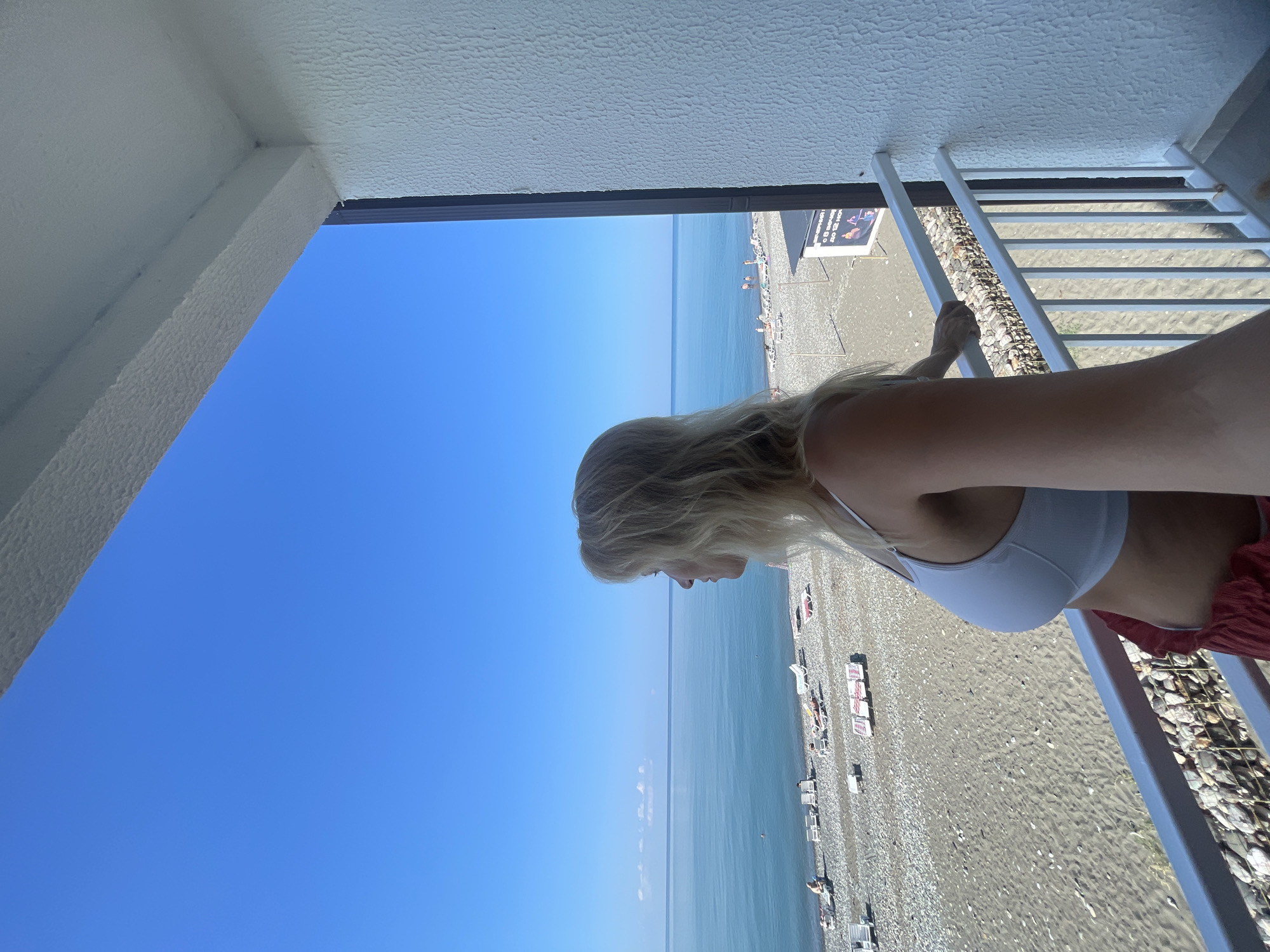 The image depicts a woman with blonde hair, wearing a white sports bra and red shorts, standing on a balcony overlooking a beach in Giachrypsh, Abkhazia. The woman is looking out at the sea, which is a calm blue. The beach is covered in pebbles and sand, with several lounge chairs and umbrellas set up. In the background, a few people can be seen on the beach. The sky is clear and blue. The balcony has a white railing and a textured white wall. The overall scene is peaceful and suggests a vacation setting.