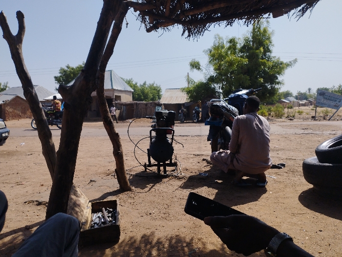The image shows a scene in Tongo, Nigeria (latitude: 10.664, longitude: 11.364). A man in a white long-sleeved shirt is crouched down, working on a motorcycle with its rear wheel off. He is positioned under a thatched roof structure. Behind him, a stack of used tires sits on the ground. To the left of the man, a large air compressor with a black tank is visible. In the distance, a man in a yellow and blue shirt rides a motorcycle away from the scene. There are various buildings in the background, including a small, white building with a blue roof. The ground is sandy and dry, with a single tree in the foreground and a few trees in the background. The image was likely captured by someone sitting on the ground, holding a black smartphone in their hand. The hand and the bottom portion of the smartphone are visible in the foreground.