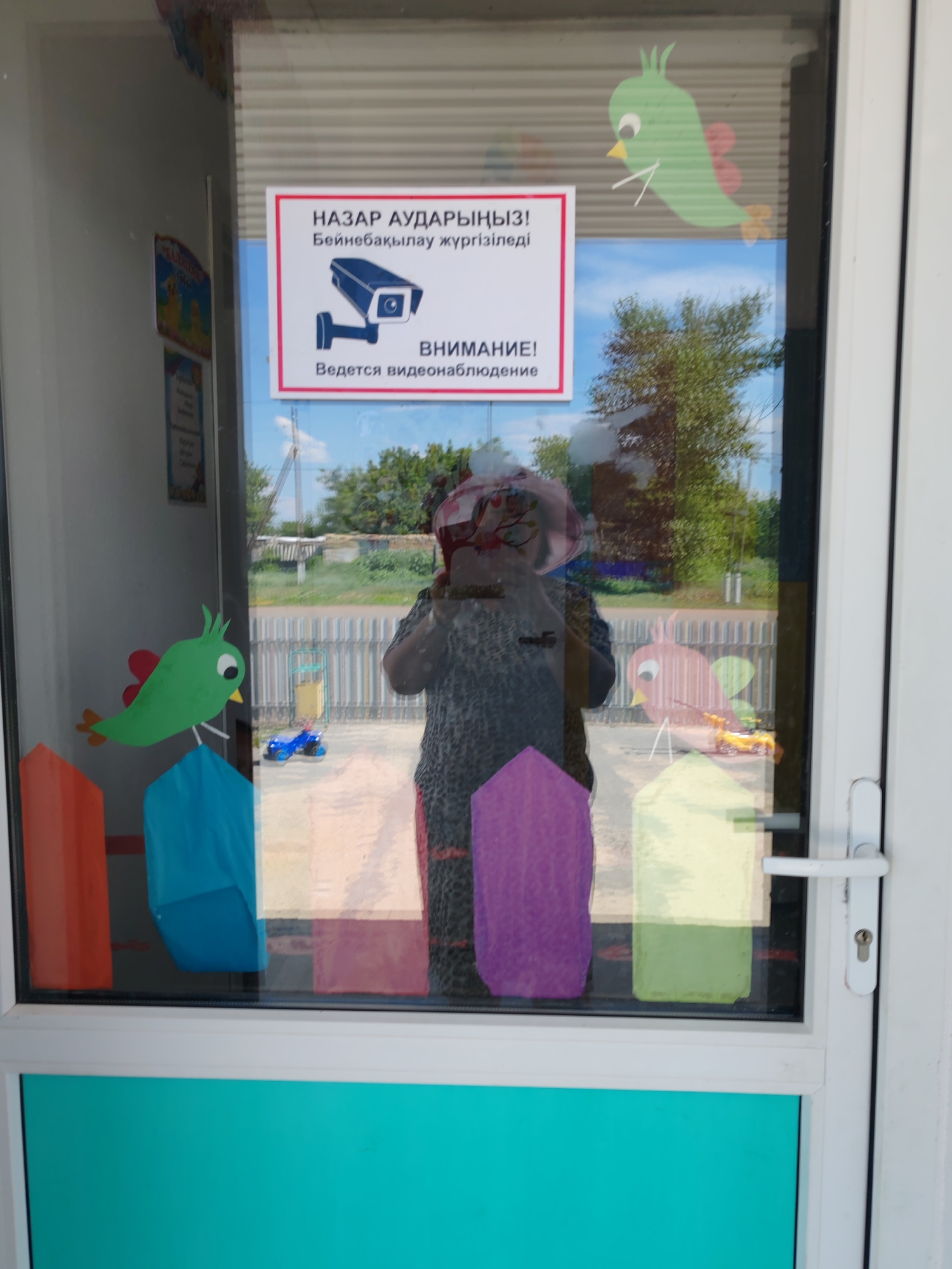 The image is a close-up shot of a glass door with a sign that reads "NAZAR AUDARYᴎᴎIЗ! Бейнебақылау жүргізіледі" in Kazakh, which translates to "ATTENTION! Video surveillance is being conducted." Below this text, there is an illustration of a surveillance camera, and further down, the text "ВНИМАНИЕ! Ведется видеонаблюдение" in Russian, meaning "ATTENTION! Video surveillance is being conducted."
The sign is framed in red and white and is affixed to the glass door. The reflection in the glass shows a person wearing a patterned headscarf and a dark patterned shirt, holding what appears to be a camera or phone, possibly taking the photo. Behind the person, there are trees and a fence, suggesting an outdoor environment.
On the glass door itself, there are colorful cutouts of birds and geometric shapes, likely decorative elements. A blue toy car is visible on the ground through the glass. The bottom portion of the door is a solid turquoise color. The location is identified as Akzhar, Kazakhstan. The scene appears to be a commercial or public building entrance, possibly a kindergarten or school given the decorative elements.