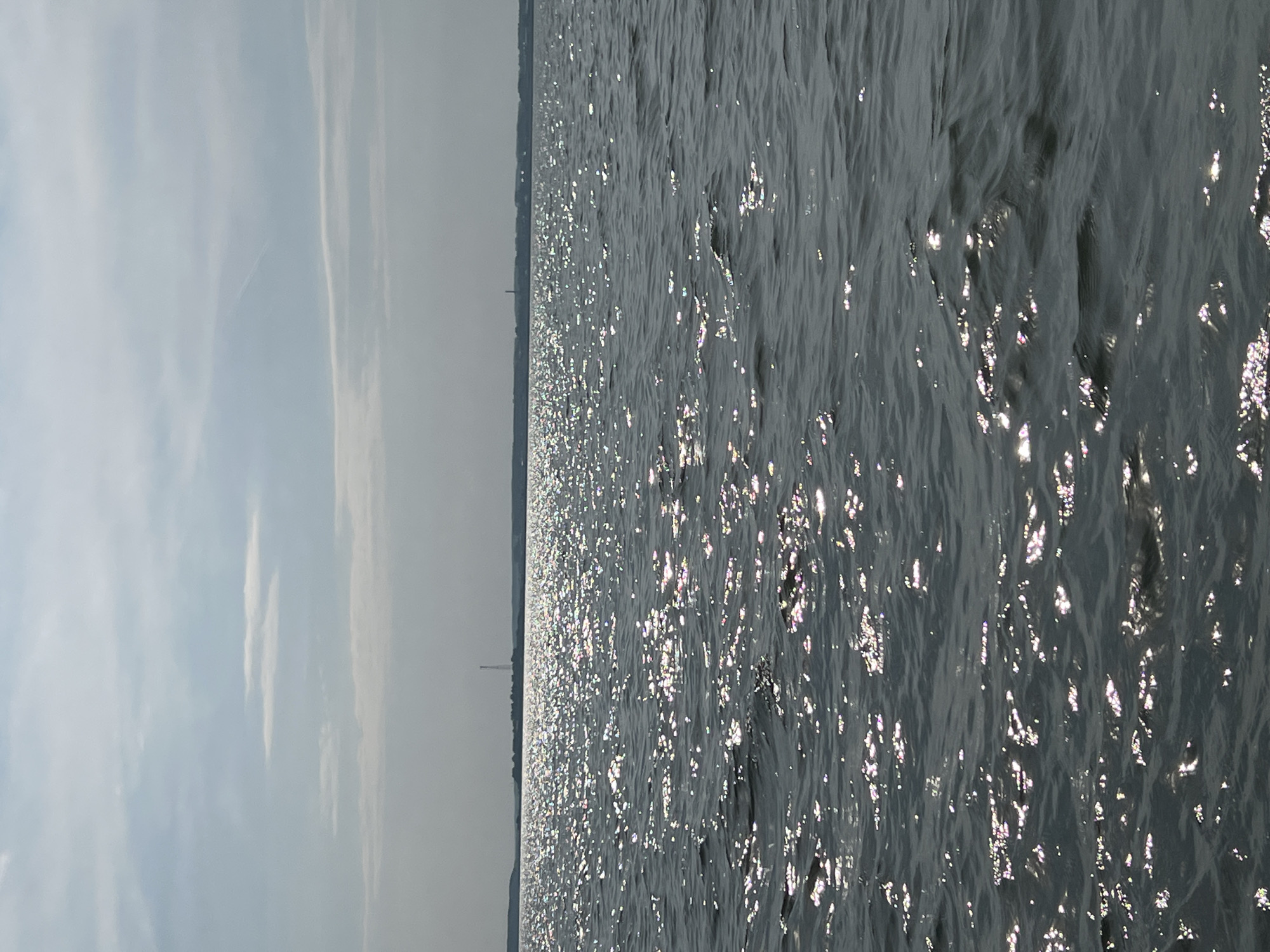 The image depicts a wide expanse of dark, choppy water under a cloudy sky. Sunlight reflects off the surface of the water, creating numerous shimmering highlights. In the distance, a hazy shoreline is visible with faint outlines of structures, possibly including a tower. The sky is overcast with scattered clouds, some appearing thin and wispy. The overall impression is one of a cool, possibly windy day on a large body of water.