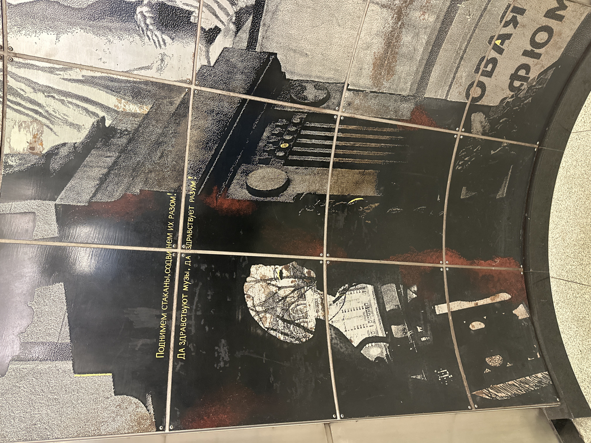 The image depicts a close-up view of a curved mural, likely located in a Moscow metro station. The mural is composed of large, dark panels, with lighter, textured elements and red accents creating a collage effect. Prominently displayed in yellow Cyrillic text are two phrases. The first reads "ПОДНИМЕМ СТАКАНЫ, СОДВИ НЕМ ИХ РАЗОМ!", which translates to "Let's raise our glasses, let's move them together!". The second phrase, "ДА ЗДРАВСТВУЮТ МУЗЫ, ДА ЗДРАВСТВУЕТ РАЗУМ!", translates to "Long live the muses, long live reason!".
The artwork features stylized imagery, including a silhouette of a person with a head formed by a textured architectural scene and foliage. Other panels show abstract textures, possibly representing buildings or urban elements. The overall impression is one of a propaganda or artistic statement, common in Soviet-era public spaces, emphasizing culture and intellect. The lighting suggests an indoor environment, consistent with a metro station. The curved surface of the mural indicates it is installed along a tunnel or arched ceiling.