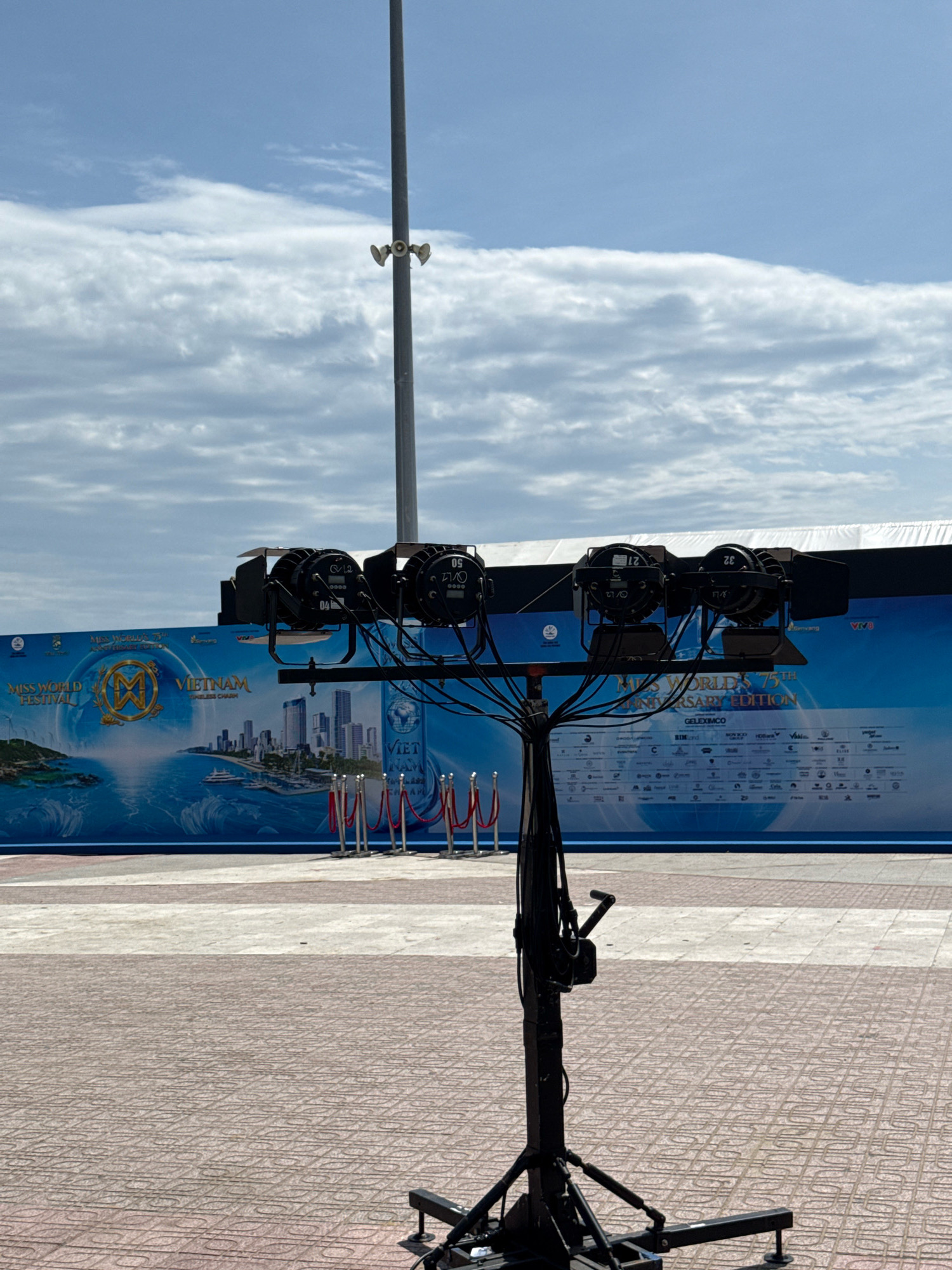 This is a photograph taken outdoors in Nha Trang, Vietnam, likely during the Miss World festival. The background features a large blue banner with text and images, possibly advertising the event and its sponsors. The banner displays a cityscape, a body of water with a ship, and the Miss World logo. In the foreground, a portable lighting rig with multiple spotlights is set up. Red velvet ropes on stanchions are visible in front of the banner, suggesting a controlled access area. The ground is paved with patterned tiles. The sky above is cloudy. There are no people in the frame.