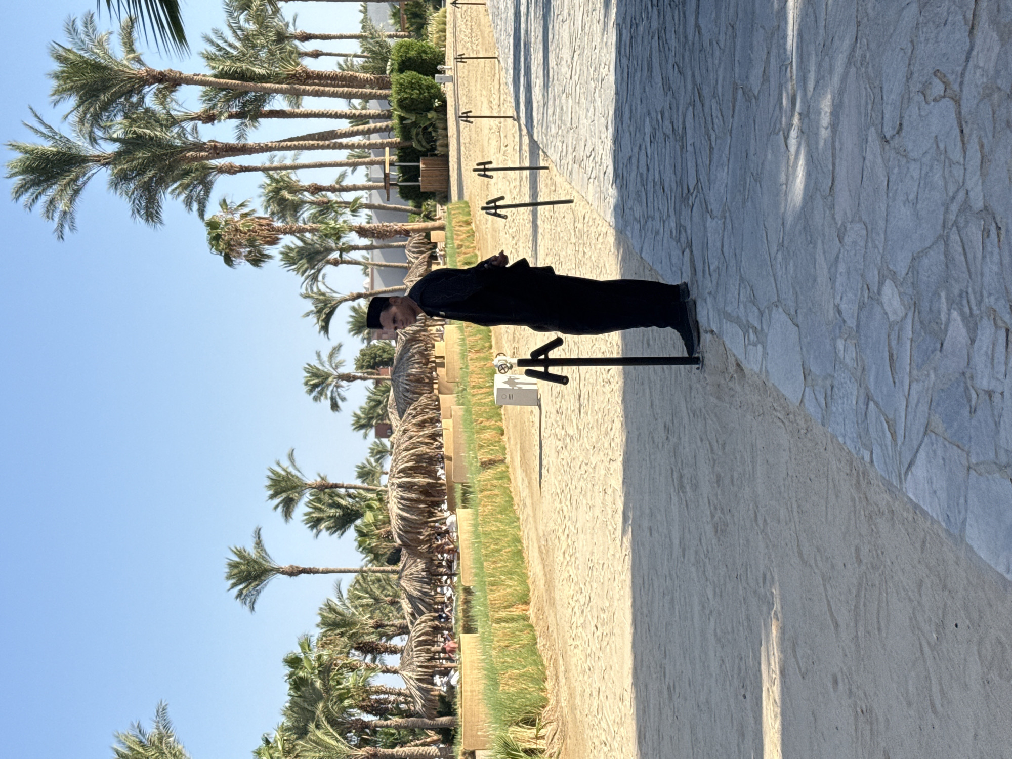 A man in a black uniform and kufi stands on a paved walkway in Hurghada, Egypt. The walkway is made of irregular light grey flagstones. To his left is a sandy area with a line of stanchions and ropes, beyond which are thatched umbrellas and palm trees. The man is looking to his left, away from the camera. The background consists of more palm trees and some buildings. The sky is clear and blue.