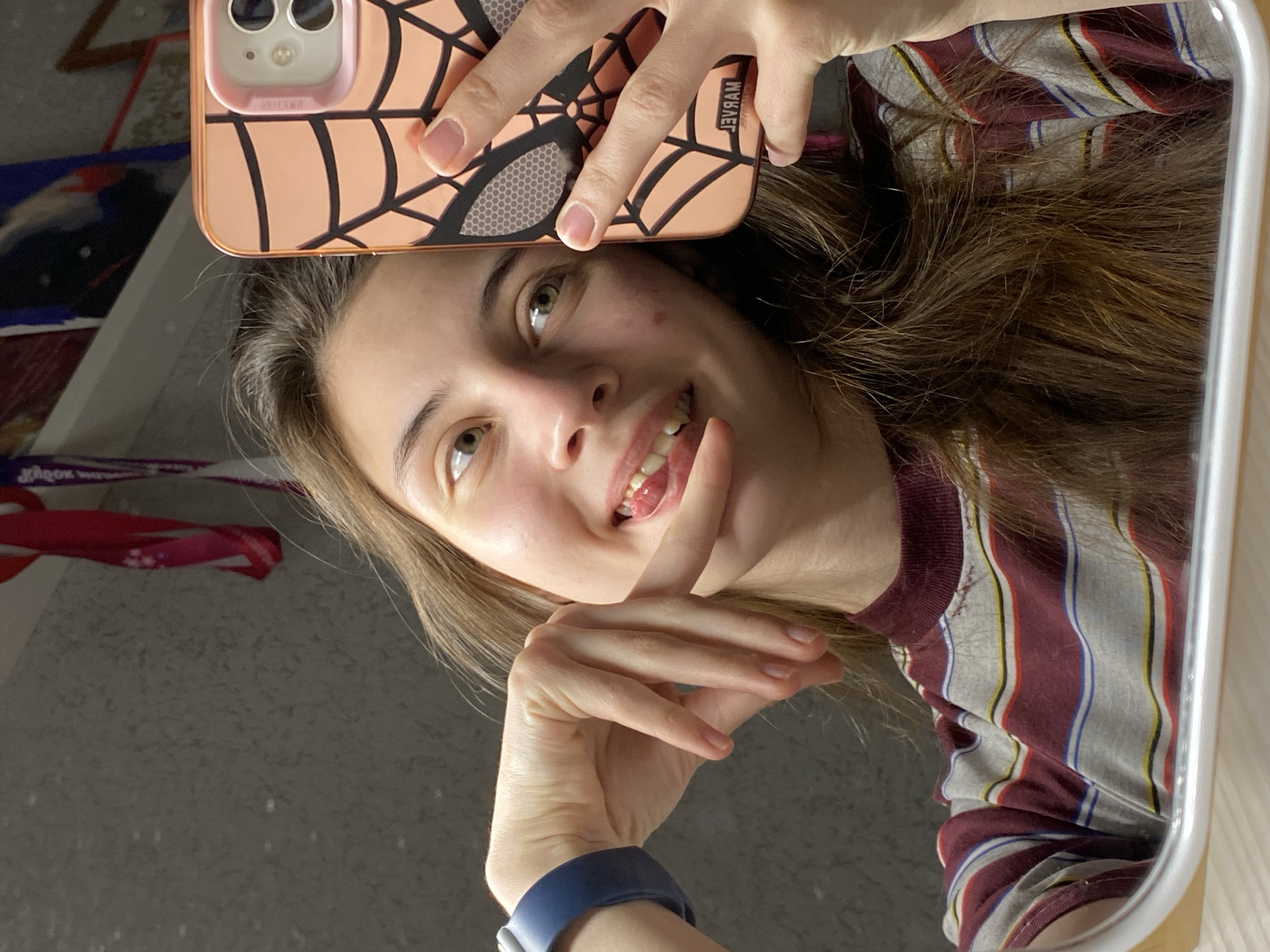 A young woman is captured in a mirror selfie, smiling with a playful expression, her tongue slightly extended, and a finger near her mouth. The image is rotated clockwise, showing her looking upwards and to the right. She has long, light brown hair and light-colored eyes. She is wearing a striped t-shirt with vertical patterns in maroon, grey, blue, yellow, and red.
She holds a smartphone, likely an iPhone, in her right hand, which is prominent at the top of the frame. The phone is encased in a peach-colored protective cover featuring a black spiderweb design. The word "MARVEL" is clearly visible on the case, appearing mirrored as "MARЯAM." Faint, mirrored text resembling "2010" or a similar alphanumeric sequence is also discernible on the case near the camera module. On her left wrist, she wears a blue wristband, possibly a smartwatch.
The setting appears to be indoors, with the woman's reflection framed by the mirror's edge on the right. The background is mostly dark and indistinct, but a textured, light grey wall is visible behind her, along with some blurry elements such as a purple ribbon-like object and a dark item with red and blue accents. Her face shows a few small blemishes. The overall mood is casual and cheerful.
This scene is located in Votkinsk, Russia.