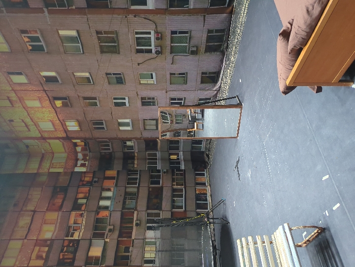 The scene depicts an indoor set designed to replicate an urban outdoor environment, likely a courtyard in Moscow, Russia, surrounded by residential buildings. A panoramic backdrop shows multi-story, grey concrete apartment buildings, characteristic of Soviet-era architecture, with numerous windows, some reflecting a warm, golden light. Visible on the building facades are exposed wiring, varied window styles, and air conditioning units. The foreground features a dark gray, flat floor marked with light-colored tape and dark stains. On the left, a light-toned slatted bench with a weathered frame is positioned, along with a large, black trampoline featuring a dark metal frame. Near the center-right, a freestanding, rectangular mirror with a wooden frame stands upright, reflecting portions of the background buildings and a small table with a chair. In the lower right, a section of a light-brown wooden bed frame with two brown pillows is visible. No individuals are present.