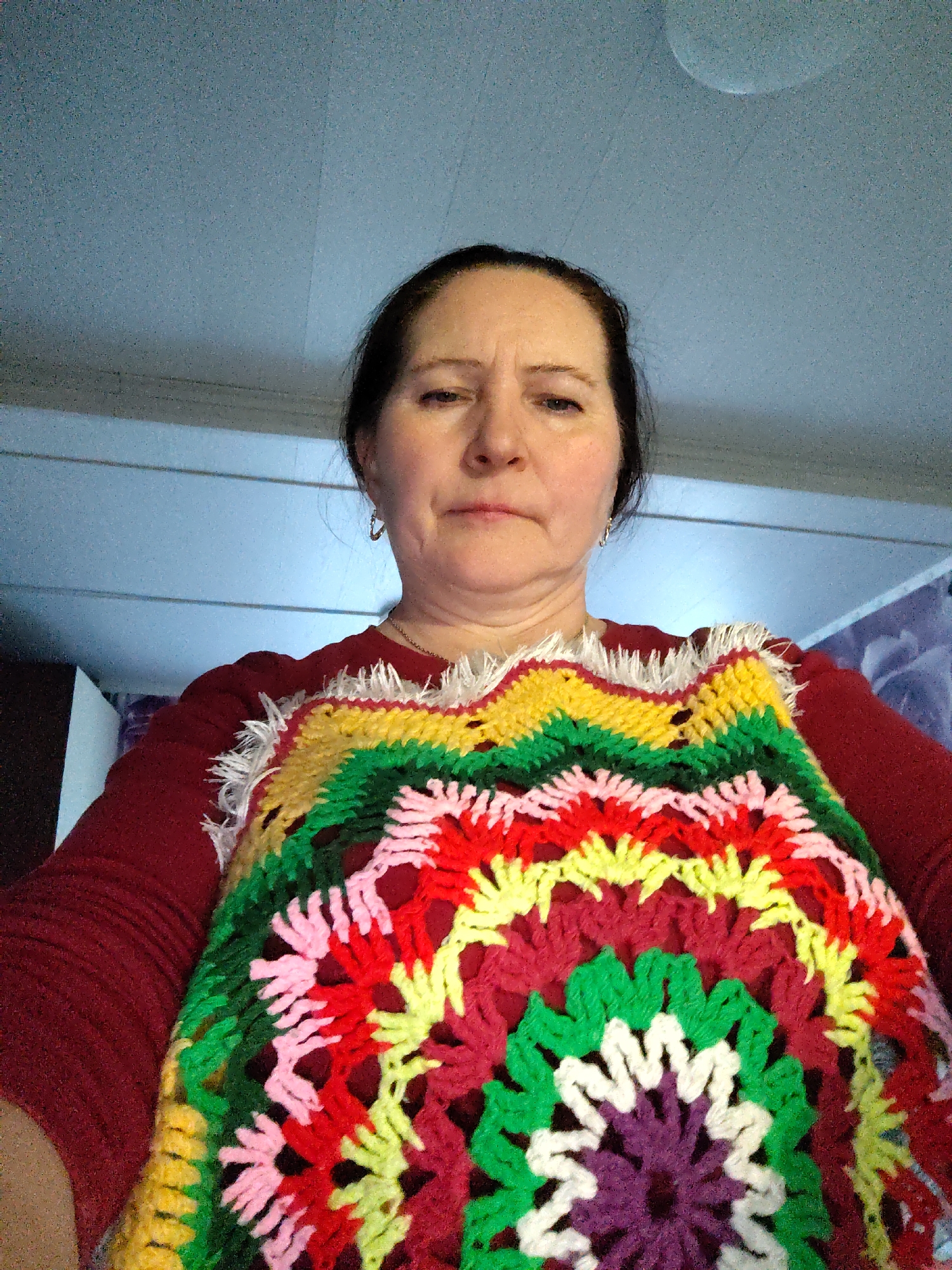 A middle-aged woman is depicted in a close-up, likely selfie-style, portrait shot indoors. She has dark hair pulled back and a neutral to slightly serious expression, looking directly at the camera. She is wearing a red long-sleeved shirt.
Over her shirt, she prominently displays a brightly colored, intricately crocheted or knitted garment or item. This piece features a large circular pattern, resembling a granny square, with a central purple motif surrounded by concentric rings of white, green, red, yellow, pink, and dark red/maroon yarns. The vibrant colors create a striking visual. The top edge of the crocheted piece has a decorative fringe of white yarn, adding texture. The woman wears small hoop earrings and a simple chain necklace.
The background shows a light blue-grey ceiling, which appears textured and has faint lines, possibly indicating structural elements or paneling, along with a barely visible circular shape that might be a light fixture. Portions of indistinct light-colored walls are also visible on either side of her head, suggesting an indoor domestic setting. The overall context is an indoor space, potentially in Сусады-Эбалак, Russia. No text is visible in the image.