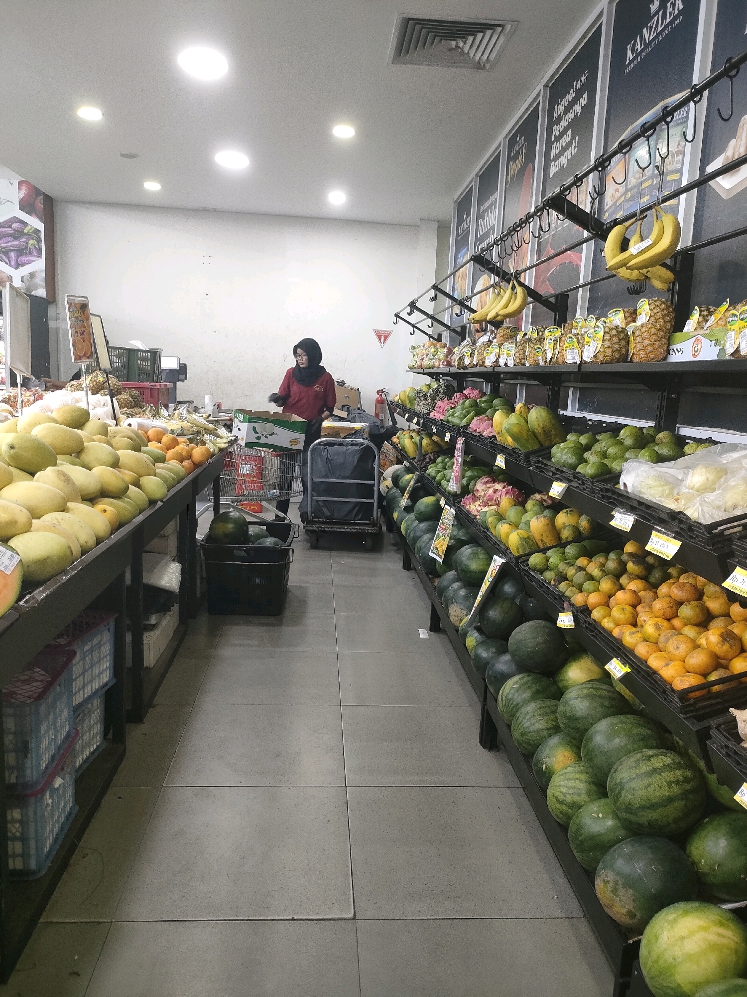The image shows the interior of a grocery store in Batam, Indonesia, displaying a wide variety of fresh produce. In the foreground, a large display of yellow mangoes is prominent. To the right, several shelves are filled with fruits including watermelons, oranges, papayas, and avocados. A woman wearing a dark headscarf and a red shirt is visible in the mid-ground, appearing to work at a checkout counter or sorting produce. She is standing behind a shopping cart, and a rolling cart with a black trash bag is next to her. The store is well-lit with ceiling lights and appears to have a tiled floor. Promotional posters are displayed on the wall in the background.