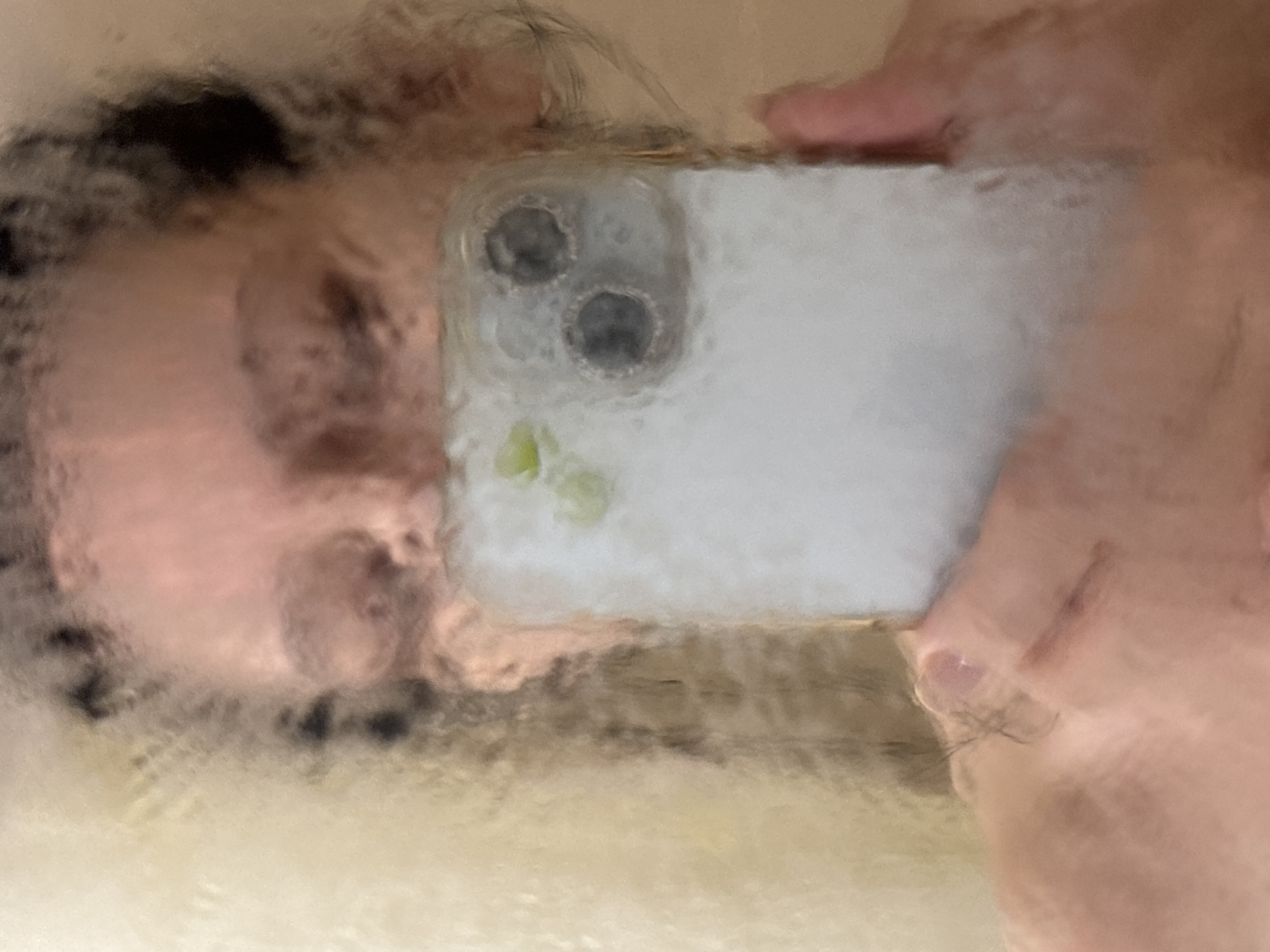 The image appears to be a blurry, close-up selfie taken through a fogged or wet surface, possibly glass or a mirror. A person's face is visible on the left side of the image, partially obscured by dark hair. The person is holding a smartphone in front of them, with the camera lens visible. The phone is a light color, possibly white or silver. The background is indistinct and appears to be a light color, potentially a wall or other surface. The location in Chaikovsky, Russia is not discernible from the visual information in the image.