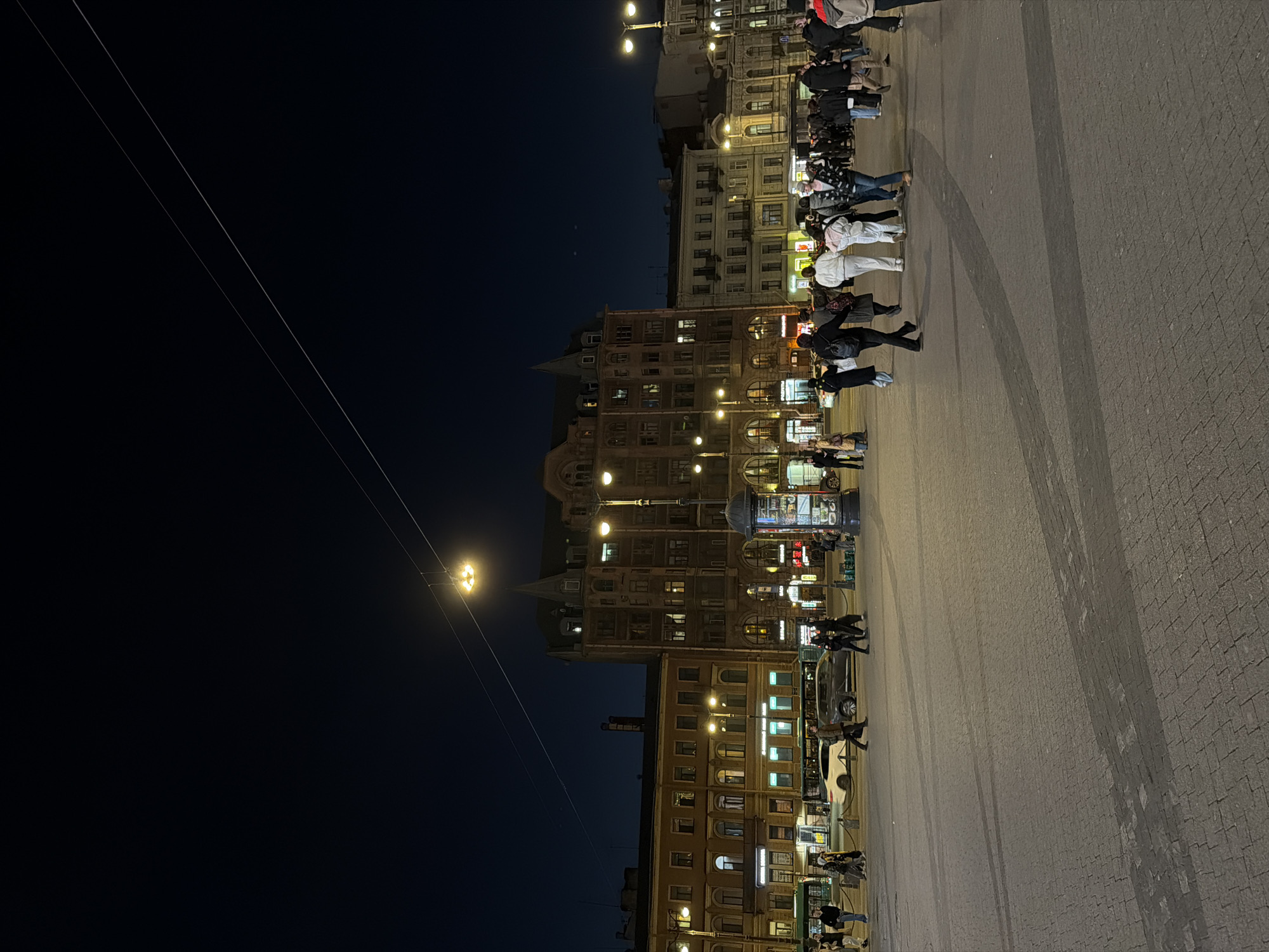 At night, a wide pedestrian plaza in Saint Petersburg, Russia, is active with numerous people walking about. Several multi-storied buildings line one side of the plaza, illuminated by internal lights and street lamps. A solitary lamp post projects light into the dark sky, and two overhead wires extend from the lamp towards the upper left of the frame. The plaza pavement has a brick or stone pattern.