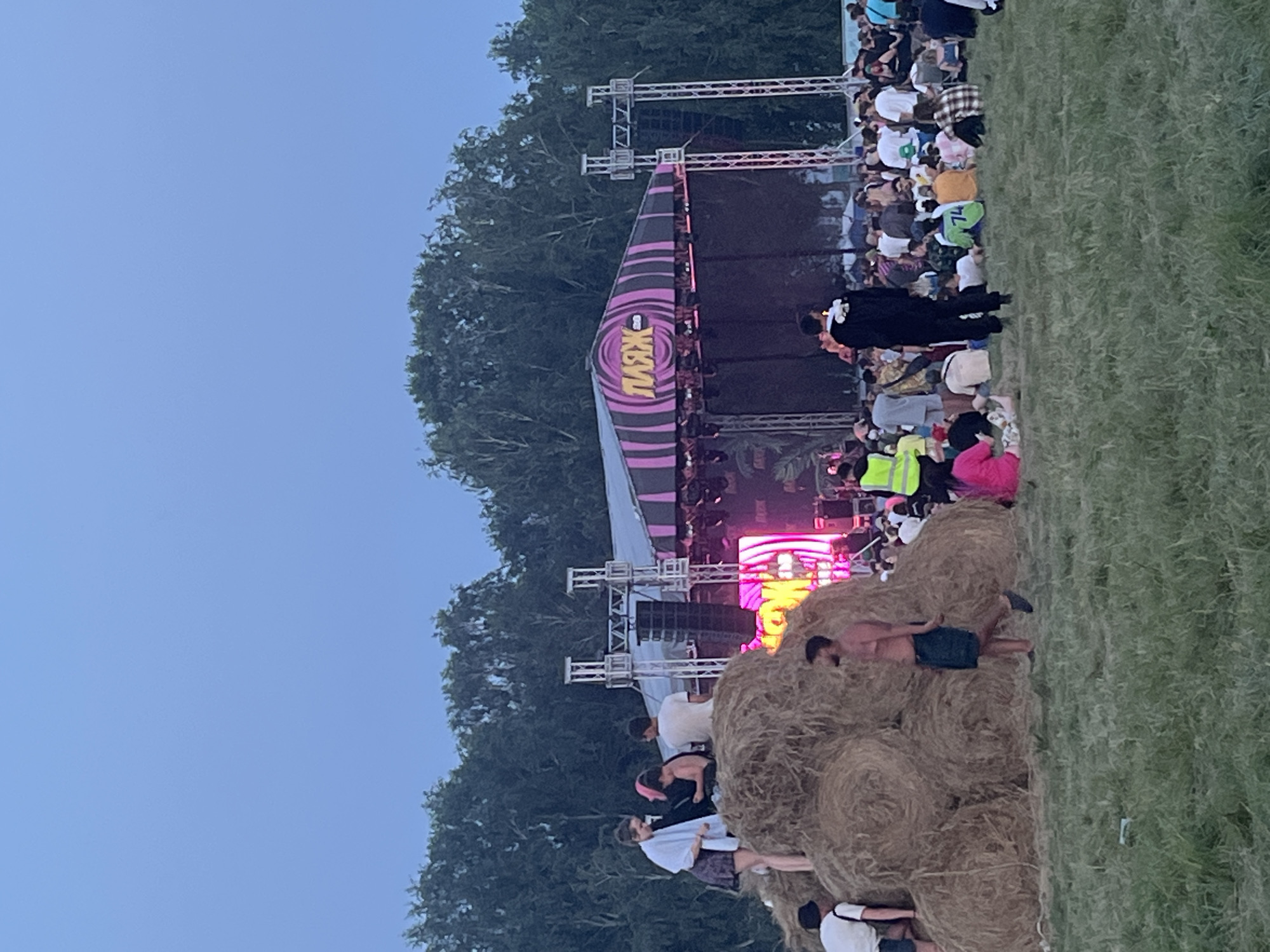 This is an outdoor event taking place in Surakovo, Russia, with a stage set up in the background. In the foreground, a large pile of hay bales serves as a seating area for some of the attendees. Several people are visible on and around the hay bales, some are sitting, some are standing, and others are climbing on them. In front of the hay bales, more people are gathered, sitting and standing on a grassy field. The stage has a pink and black striped canopy with the word "ПЛЯЖ" (Plage) written in large, yellow letters. Pink and purple lights are illuminating the stage. Trees form a backdrop behind the stage and the event area. The sky is a clear, muted blue, indicating it is either early morning or late evening. The overall scene depicts a lively outdoor festival or concert.
