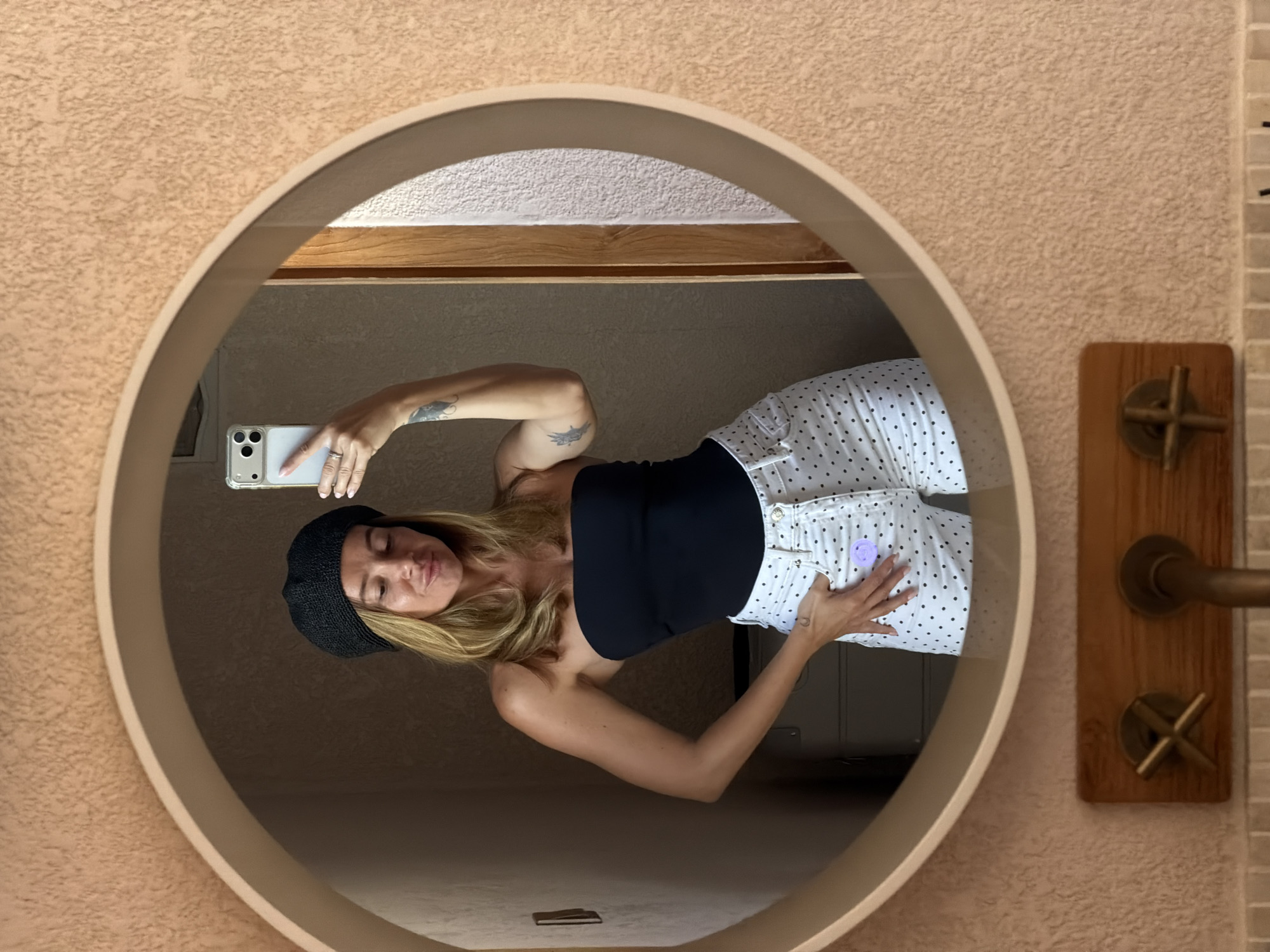 In a bathroom with textured peach-colored walls, a woman is taking a mirror selfie. She is wearing a black strapless top and white pants with black polka dots. A black beret sits on her head, and her blonde hair is down. Her left hand is on her hip, and her right hand holds up a smartphone. The mirror is circular and framed in a light-colored material. Below the mirror, a wooden block with brass faucet handles is visible against a tiled backsplash. The background reflected in the mirror shows a doorframe and part of a suitcase. The scene is set in a bathroom in Mengwi, Indonesia.