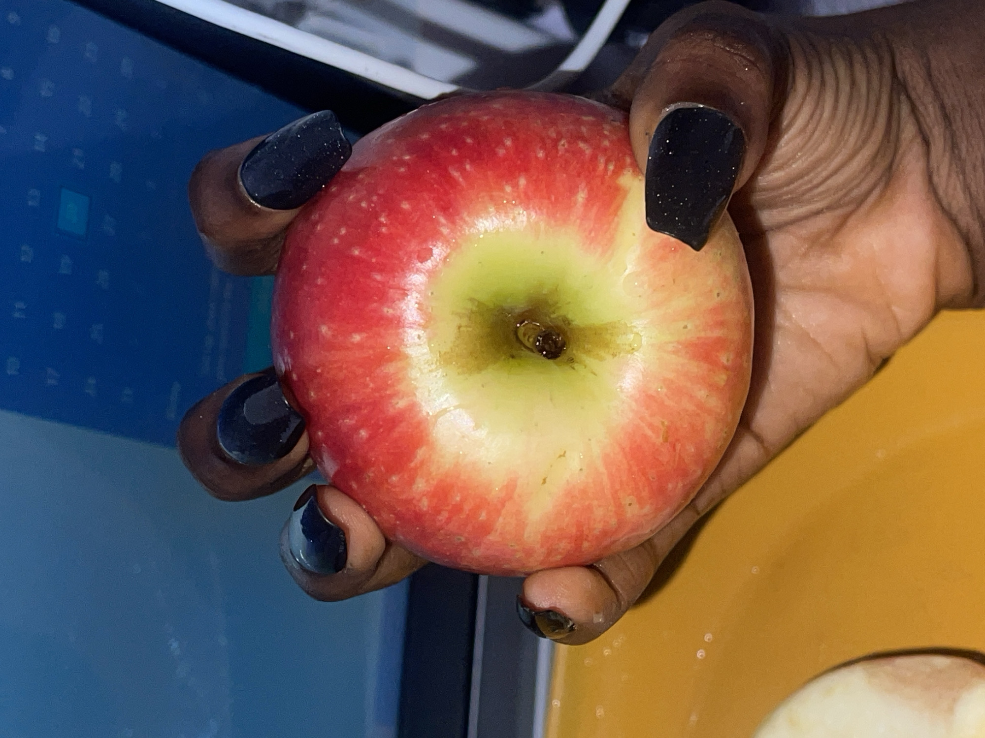A close-up shot shows a person with dark skin holding a red and green apple. The person's nails are painted black with a glittery finish. The apple has a yellowish-green center. Part of a blue screen is visible in the background on the left, and a yellow surface is on the right. The location is not discernible as being in Ado, Nigeria.