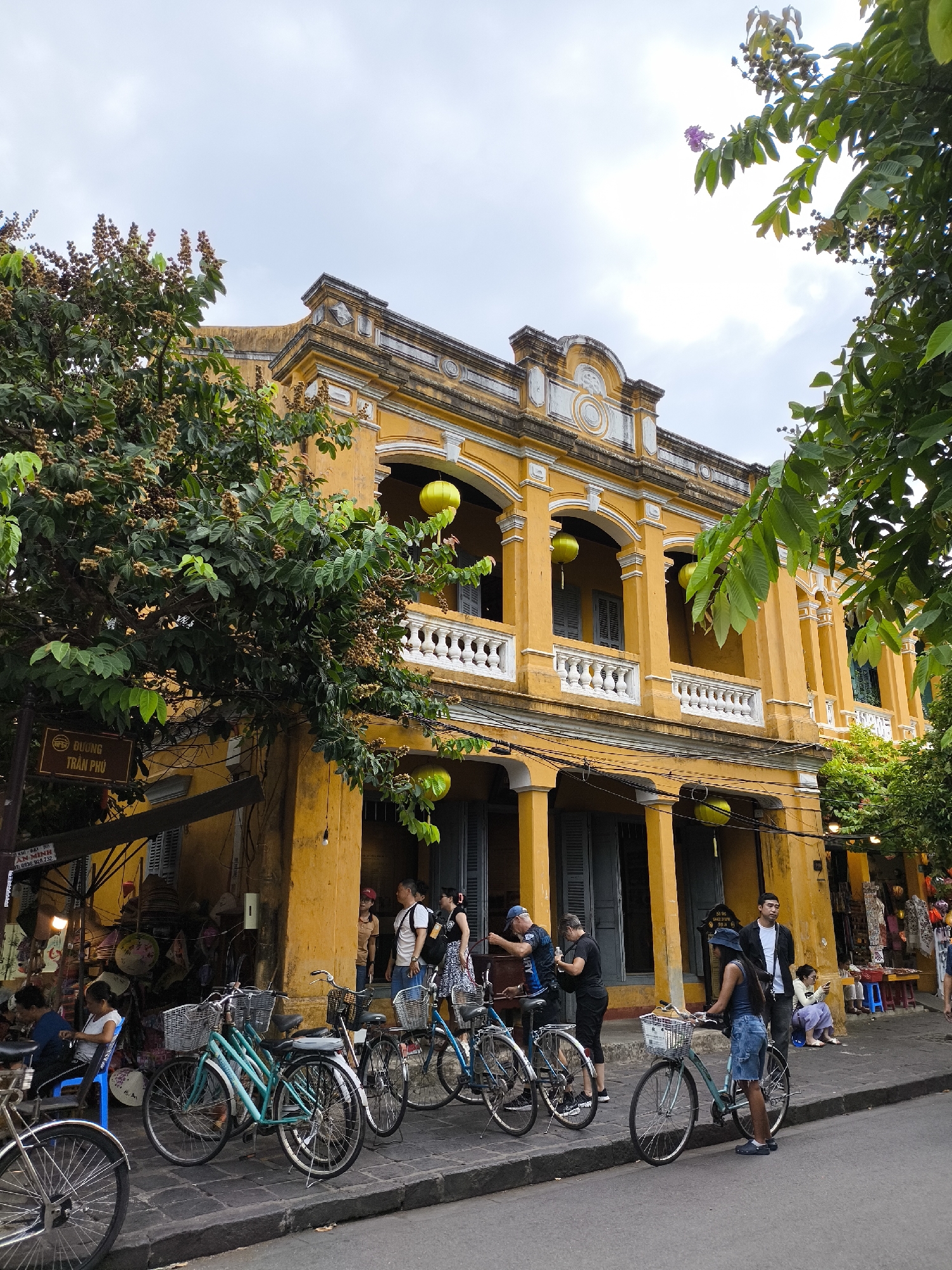 This is a street-level, eye-level, medium shot taken in Hoi An, Vietnam. The scene is dominated by a bright yellow, two-story building with classical architectural elements, including arched windows and a decorative facade. Several green lanterns are visible hanging from the building's balconies. In front of the building, a sidewalk is lined with parked bicycles, some with baskets.
On the sidewalk, there are several people. On the left, a woman sits on a blue chair near a market stall displaying woven goods. To her right, several people are standing and interacting, including a man and woman with backpacks. Further to the right, a woman in a denim skirt stands by a bicycle, and a man in a black jacket stands next to her. A woman sits on a low stool in the background. There are also various market stalls with wares visible. Trees with green leaves and some brown seed pods overhang parts of the scene. The street is paved with dark stone, and a grey curb separates it from the sidewalk. The sky is overcast.