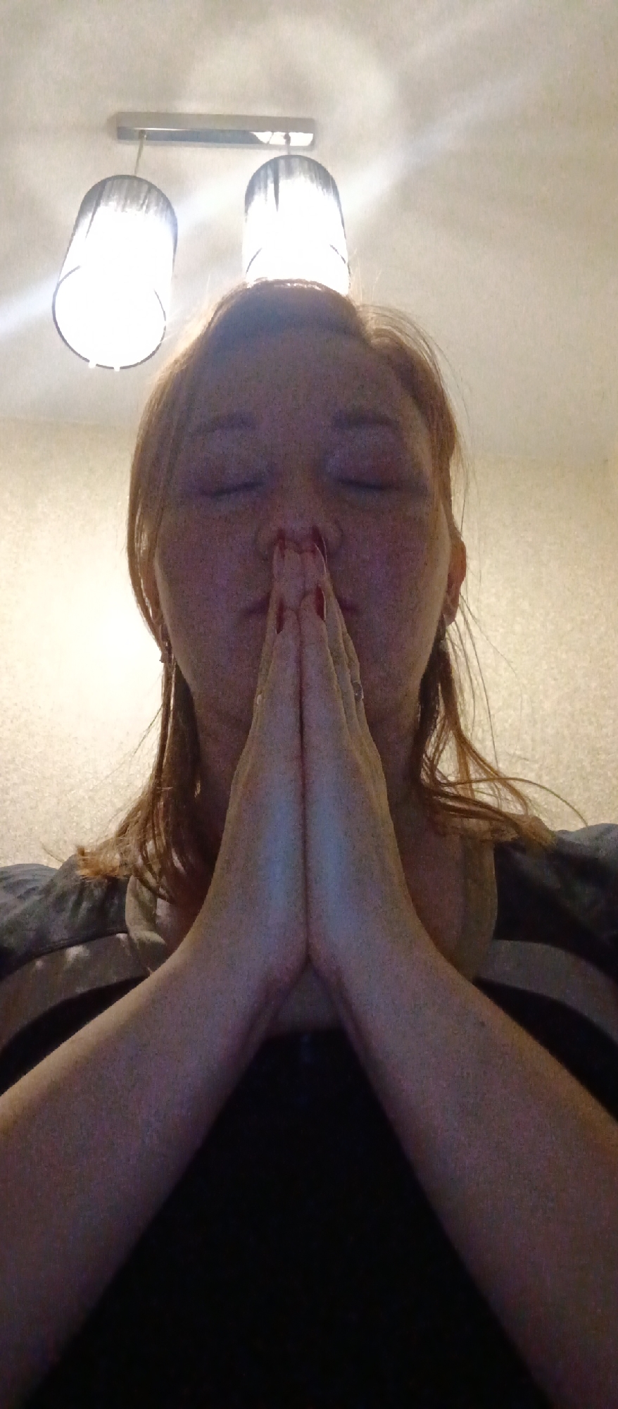 A woman with reddish-brown hair is positioned with her hands clasped in front of her nose, eyes closed. She is wearing a dark, striped shirt. Above her head is a ceiling light fixture with two lamps. The background is a plain, light-colored wall. The location and city are Unknown, Unknown.