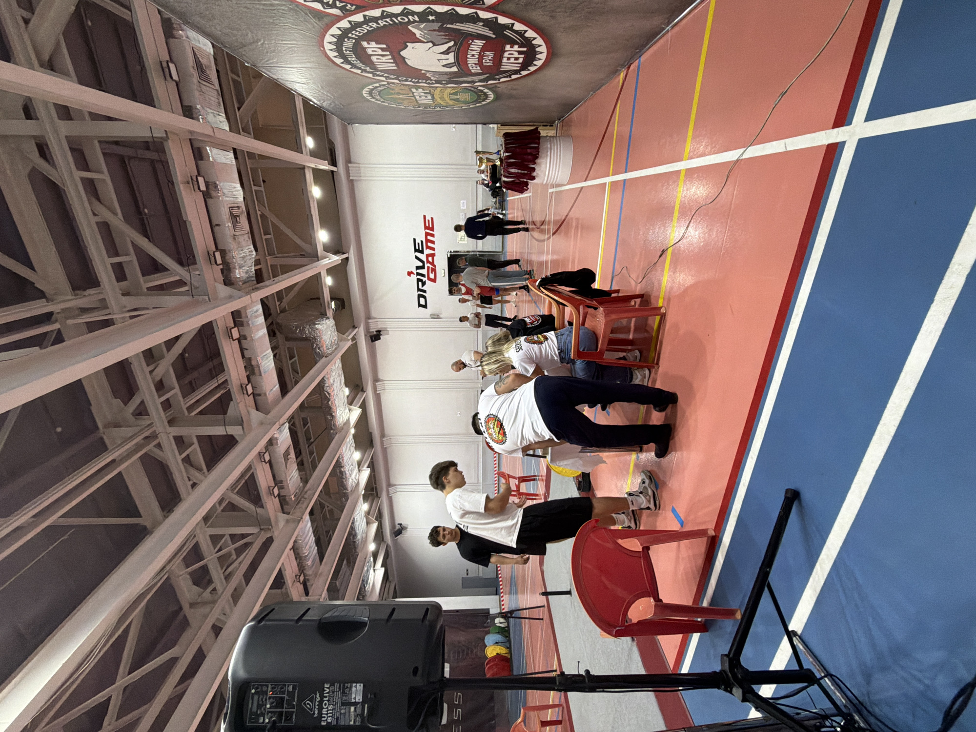 The image shows an indoor sporting event in Perm, Russia, based on the Cyrillic text “Пермский край” which means “Perm Krai” (region). A banner at the end of the room contains the acronyms "WRPF" and "WEPF," possibly related to powerlifting federations. A few people are standing, some are sitting on red chairs, and others are walking towards a raised platform or table at the far end of the room. The floor has painted lines marking different sections in red, white, yellow, and blue. The building has a high ceiling with exposed structural beams. A black speaker labeled "Behringer EUROLIVE B115D" is in the foreground. The word "DRIVE GAME" is displayed prominently on the wall.