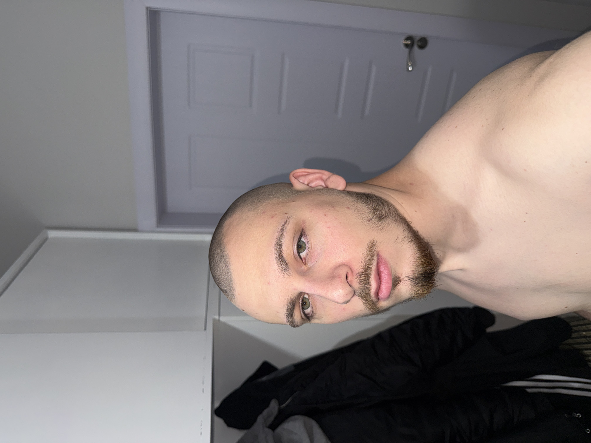 A portrait-oriented image, rotated 90 degrees counter-clockwise, depicts a young adult man indoors in Stara Zagora, Bulgaria. The subject is captured from the chest up, shirtless, with a buzzed haircut and a well-trimmed goatee beard. He has light skin, greenish-hazel eyes, and a neutral to slightly serious expression as he looks directly at the camera. His right ear is clearly visible.
In the background, a light grey or white door with two rectangular recessed panels is prominent, featuring a metallic door handle on the right side. To the left of the door, a plain light-colored wall or part of a wardrobe structure is visible. A dark, possibly black, jacket with three distinct white stripes (suggesting a sports brand like Adidas) is draped in the lower right portion of the frame, partially obscuring other clothing items.
The lighting appears to be artificial or low natural light, creating soft shadows and highlighting the subject's features. The setting is an interior room, likely a bedroom or living space, given the presence of the door and wardrobe-like structure. There is no visible text in the image.