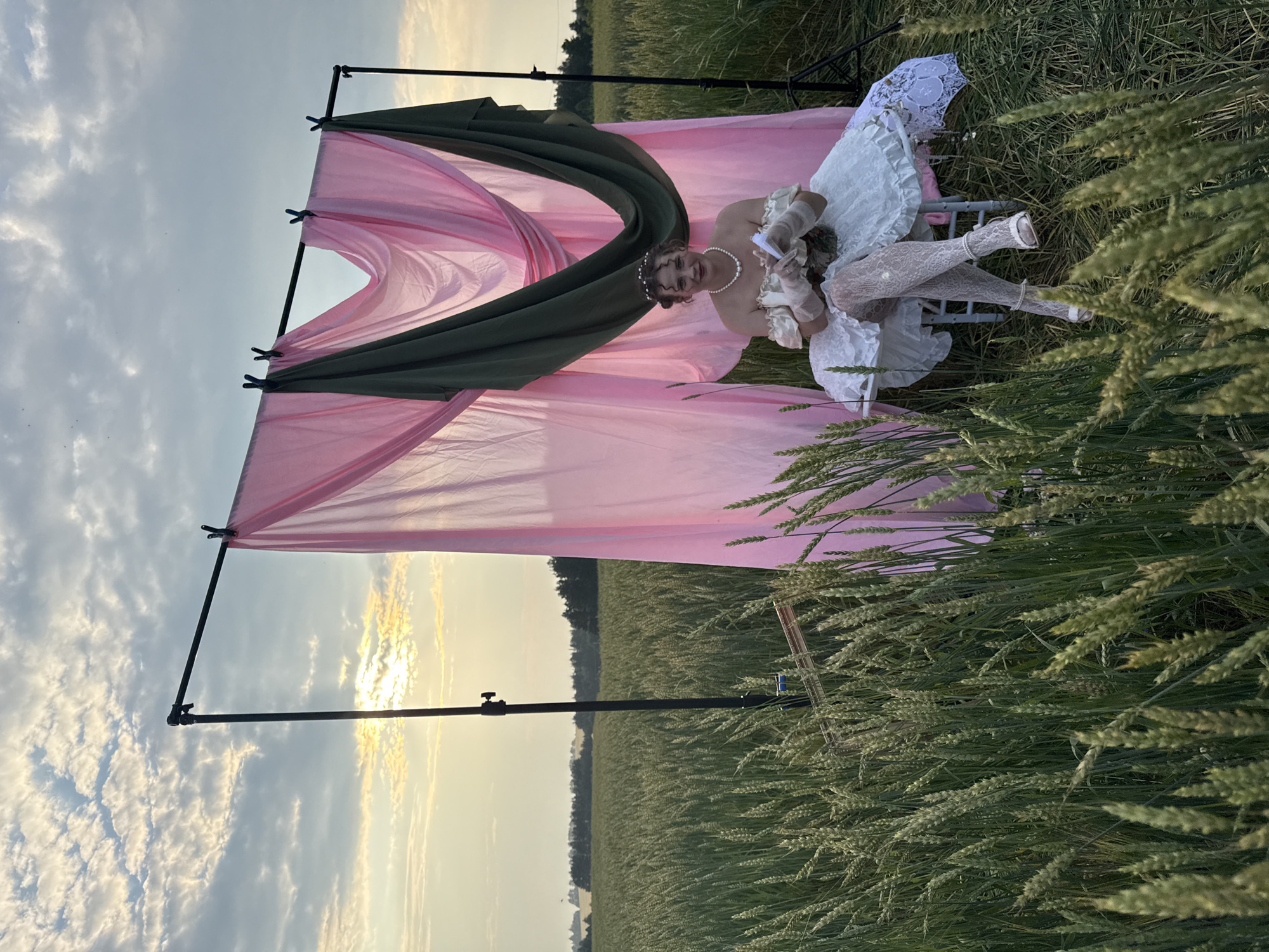 A woman in a white, ruffled dress and white lace stockings sits on a light-colored chair in a field of wheat. She is wearing a pearl necklace and has a circlet with pearls in her hair. Her arms are extended, and she holds what appear to be papers in her hands. A large, flowing pink fabric is draped over a metal frame behind her, with a dark green fabric also attached to the frame. The scene is outdoors, with a field of wheat stretching into the distance. The sky is cloudy, and the sun appears to be setting or rising, casting a warm glow. The location is identified as Malyi Bukor, Russia. The overall impression is of a staged photoshoot.