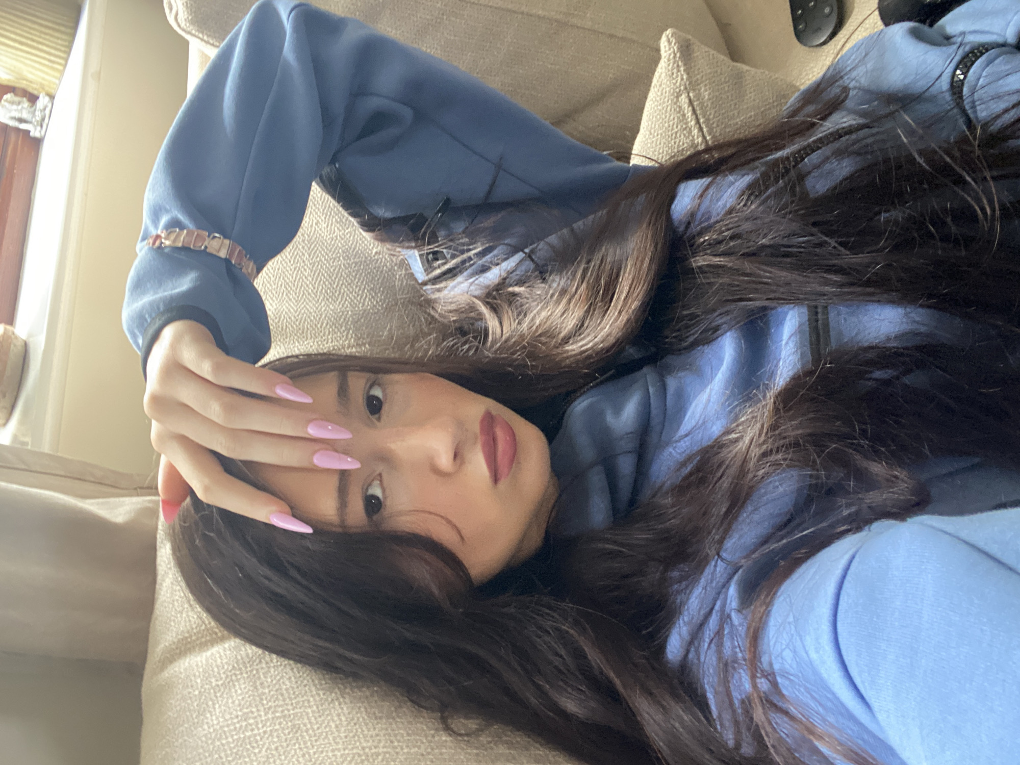 A woman is reclining on a beige sofa, wearing a light blue zip-up hooded sweatshirt. She has long, dark brown hair and pink, stiletto-shaped fingernails on the hand raised to her forehead. She is wearing a gold bracelet on the wrist of her raised hand and is looking directly at the camera. The background consists of part of a window and a neutral wall. The location is Reigate and Banstead, United Kingdom.