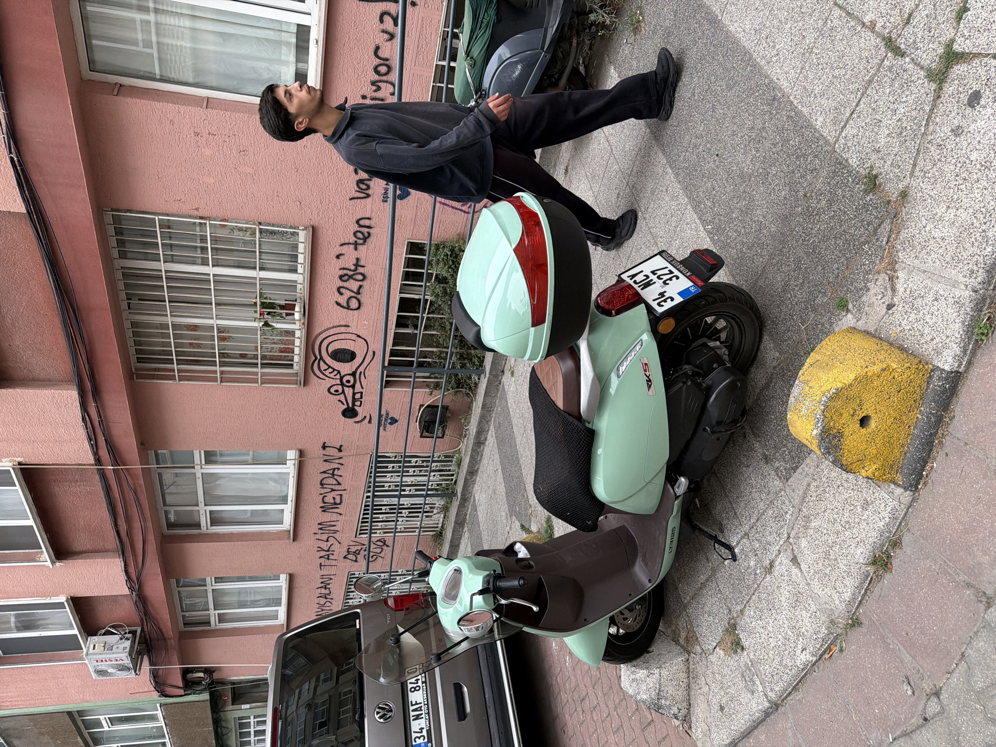In Kadıköy, Turkey, a pale pink building with white-trimmed windows dominates the background. Graffiti covers the lower portion of the building's facade, including what appears to be Turkish text and a cartoonish drawing of a mouse. A young man, dressed in a dark gray long-sleeved shirt and black pants, walks across a speckled gray concrete sidewalk. To his left, a light mint green and brown scooter is parked, facing left, with a detached mint green storage box on the rear. The scooter has a black seat cover and a license plate reading "34 NCY 327". Adjacent to the scooter is a grey car, with its license plate visible as "34 NAF 84". A yellow concrete bollard, marked with black stripes, is situated at the edge of the sidewalk in the foreground. The environment suggests an urban street scene.