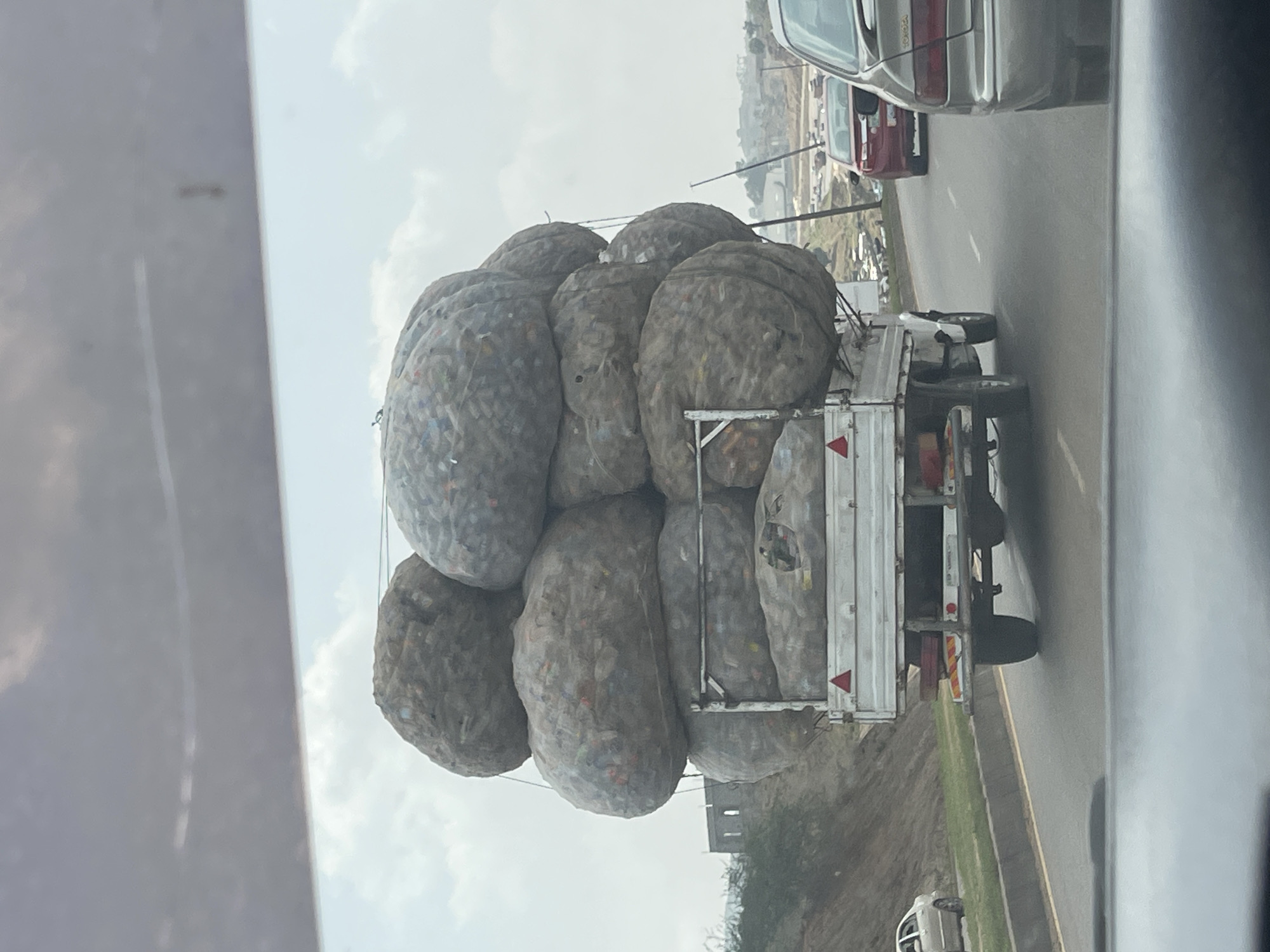 A truck is heavily laden with sacks of material, possibly waste, on a road in Kugbo, Nigeria. The sacks are bulging and appear to be secured on the truck bed. The truck is on a paved road with other vehicles nearby. The environment appears urban with buildings and vegetation visible. The image is taken from inside another vehicle, looking out the side window.