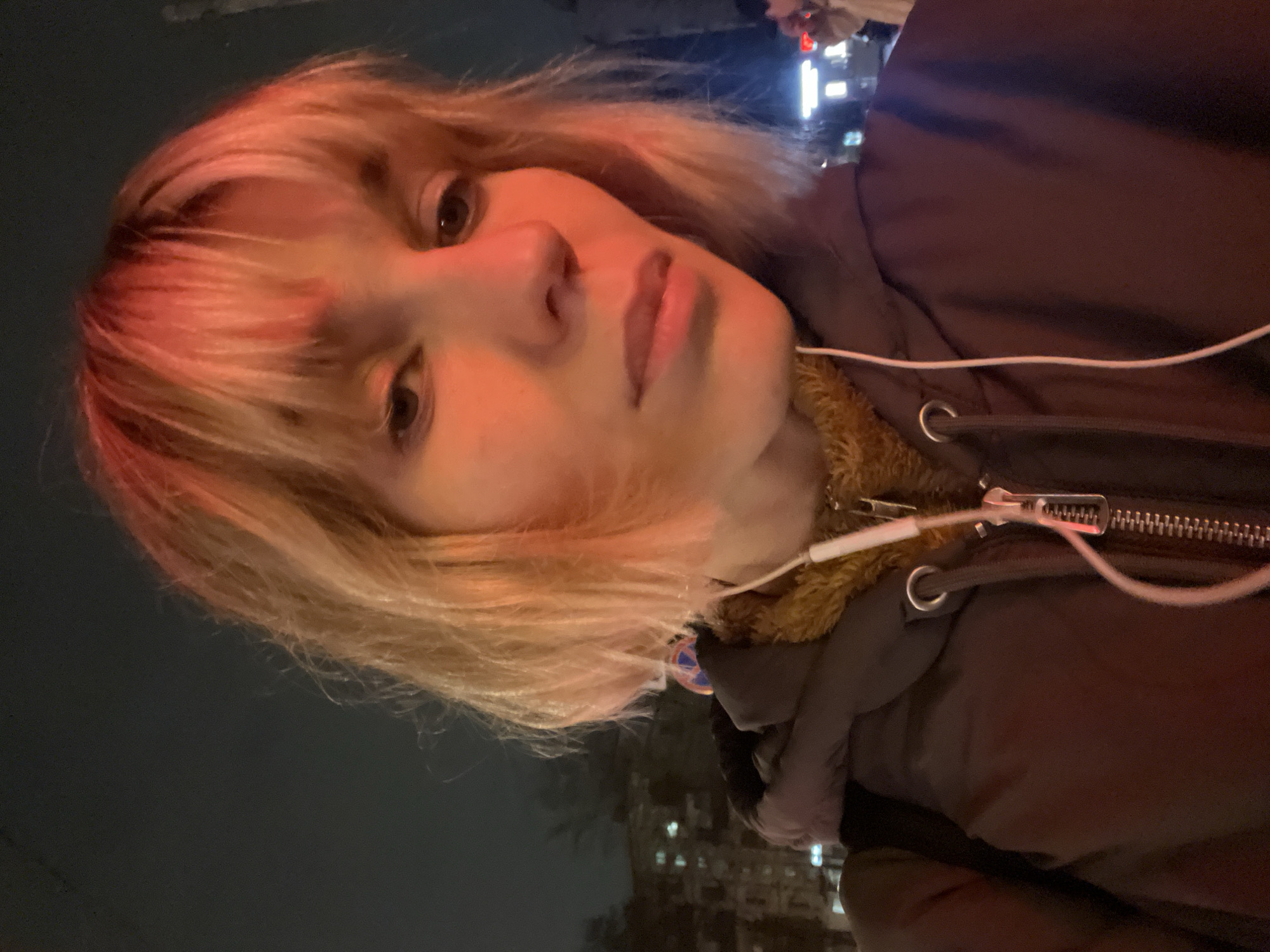 A close-up shot of a person with blonde hair and fair skin, appearing to be taken at night in an urban environment. The person is wearing a dark jacket with a fur-lined collar and white wired headphones. The background is dark, with indistinct building lights suggesting a city setting. Moscow, Russia is mentioned as the possible location for this urban setting.