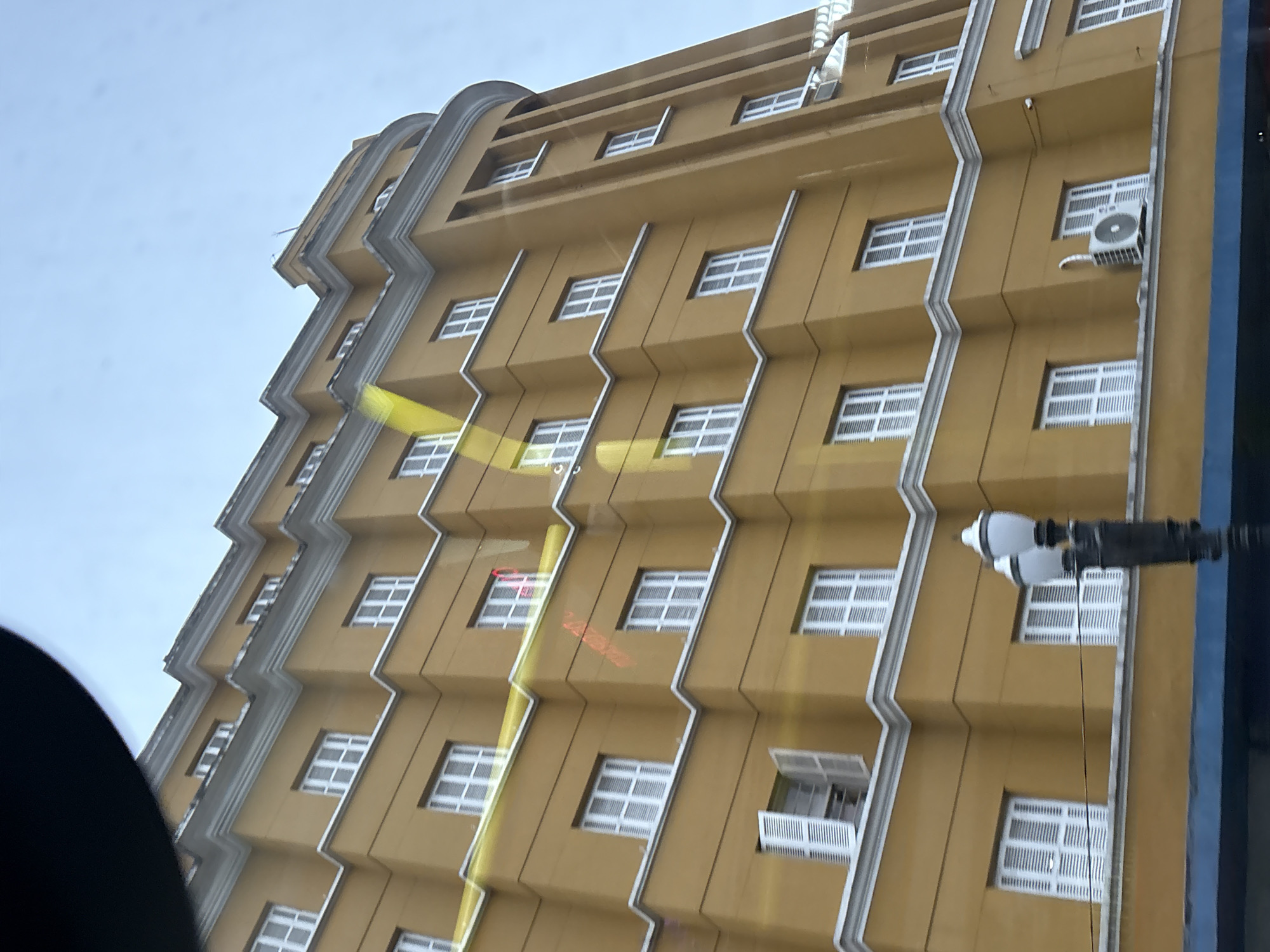 The image shows a tall, yellow building with a distinctive stepped facade and white-trimmed windows. The building is likely located in São Paulo, Brazil, given the architectural style and presence of white-shuttered windows and air conditioning units commonly found in the region. The sky is overcast, suggesting a cloudy day. A black, circular object is visible in the upper left corner, possibly a reflection or part of the camera's lens. The building's facade is punctuated by numerous windows, many of which are closed and appear to have white shutters. One window on a lower level is open, revealing a glimpse of the interior. A street lamp with a white, ornate top stands in the foreground. Yellow streaks of light or reflections are also visible across the building's surface, possibly from streetlights or vehicle headlights.
